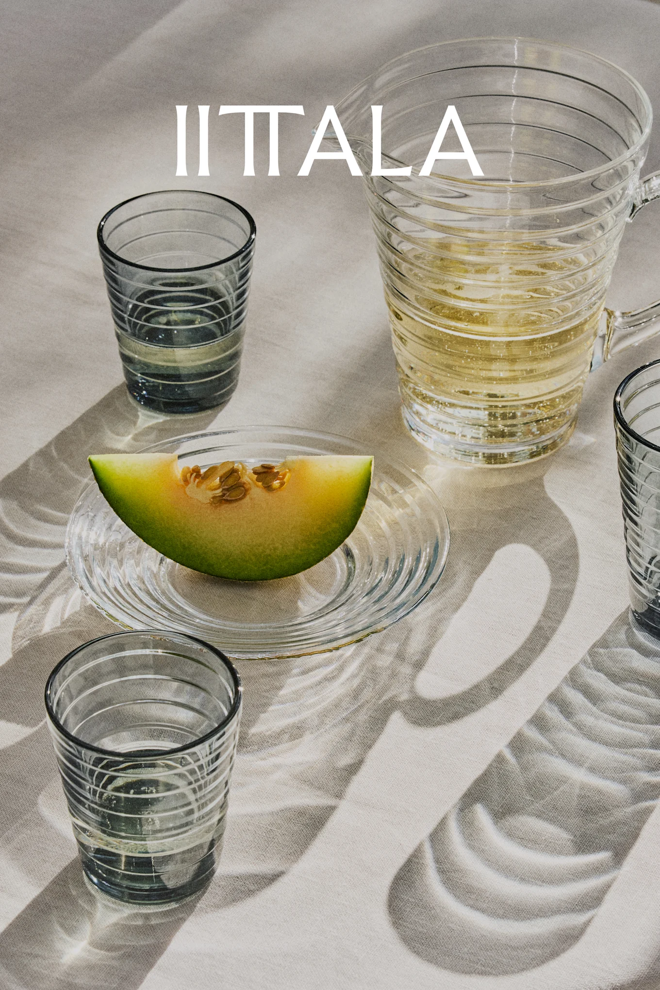 Logo IITTALA au-dessus de verres, une carafe de boisson pétillante et une tranche de melon sur une assiette, sur un tissu clair.