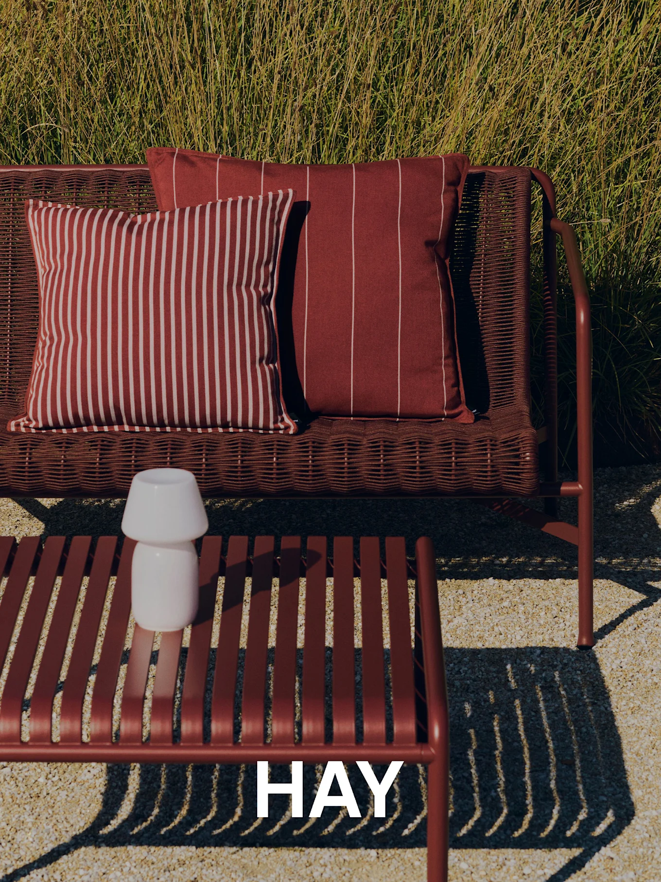 Scène d'extérieur avec un canapé rouge tressé, des coussins rayés, une table basse à lattes avec une lampe blanche et de hautes herbes en arrière-plan. Logo HAY.