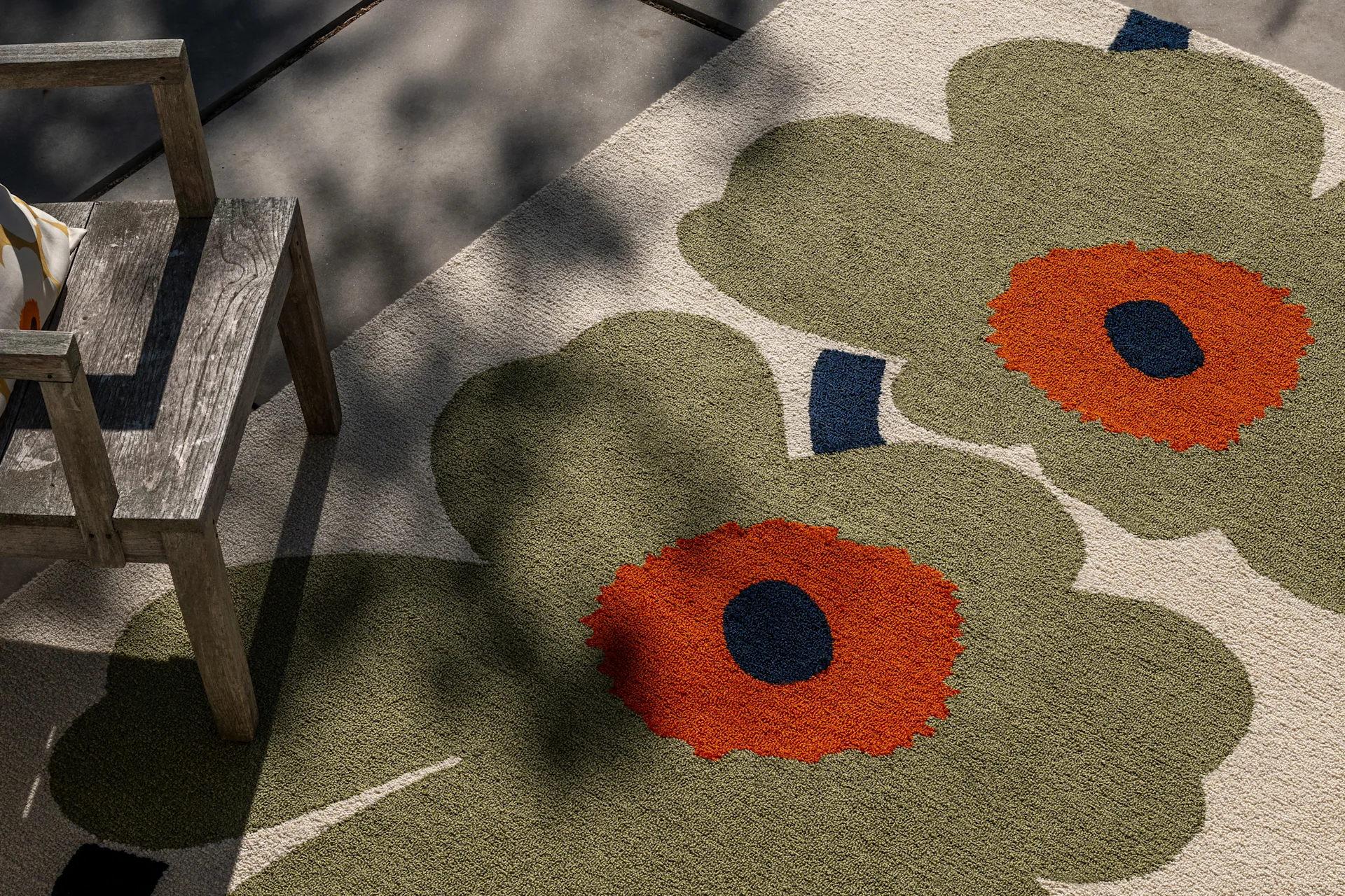 Tapis d'extérieur à grand motif floral vert olive, orange et bleu foncé, à côté d'une chaise en bois rustique sous la lumière du soleil.