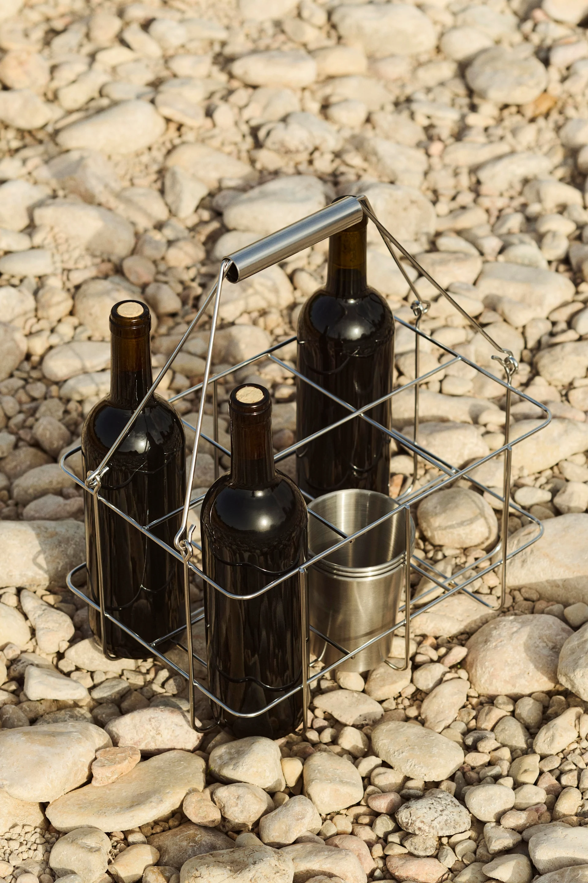 Trois bouteilles de vin et une tasse en métal dans un porte-bouteilles sur un lit de galets.