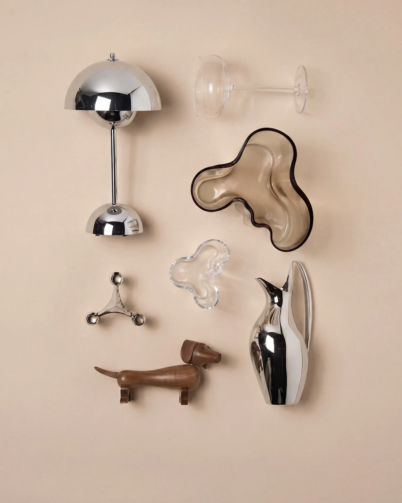 Un assortiment d'objets décoratifs: une lampe chromée, verre à vin, deux bols organiques, un candélabre, un teckel en bois et une carafe.