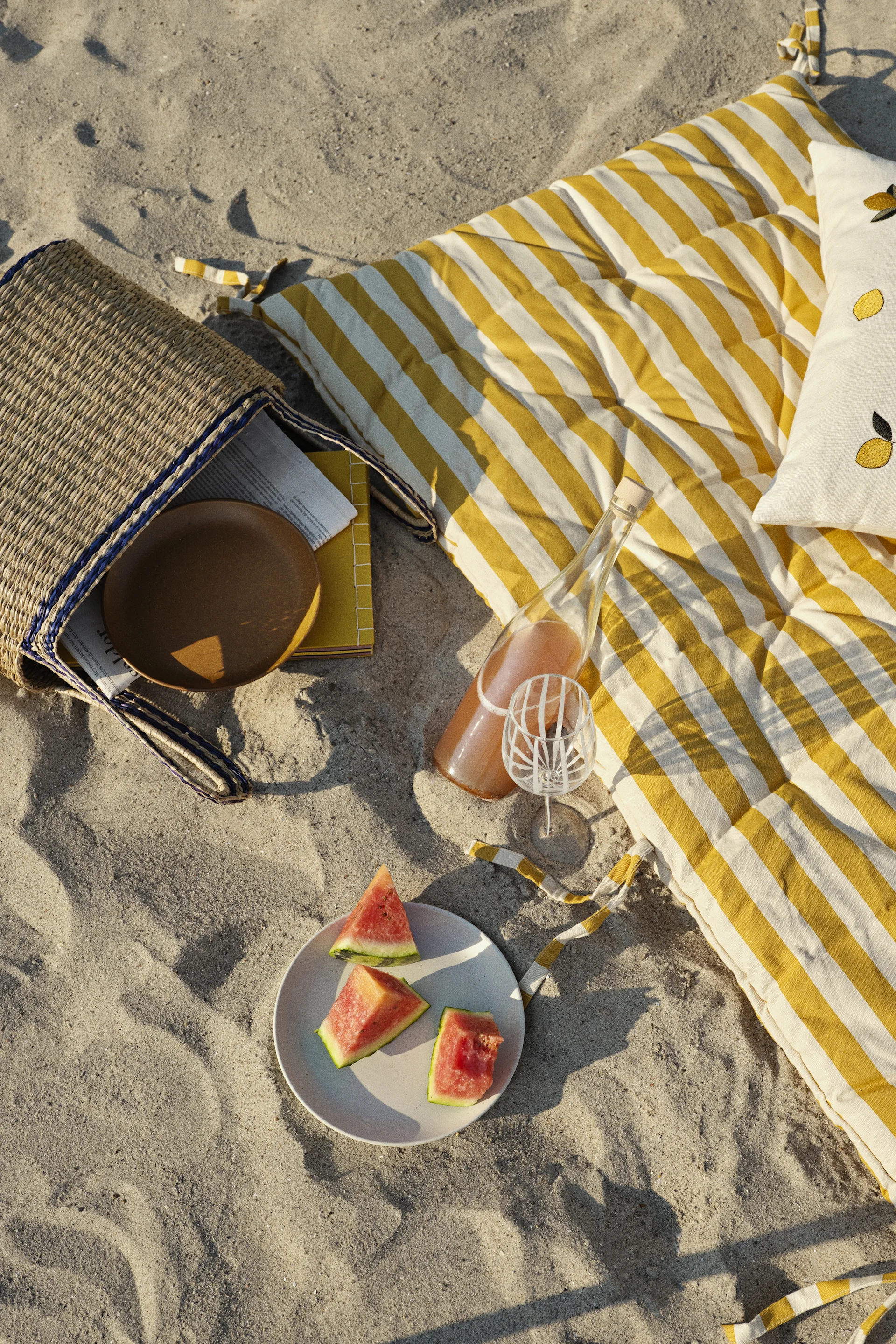 Pique-nique à la plage avec tapis rayé jaune, sac en paille, pastèque, bouteille de boisson, verre à motifs et coussin citron.