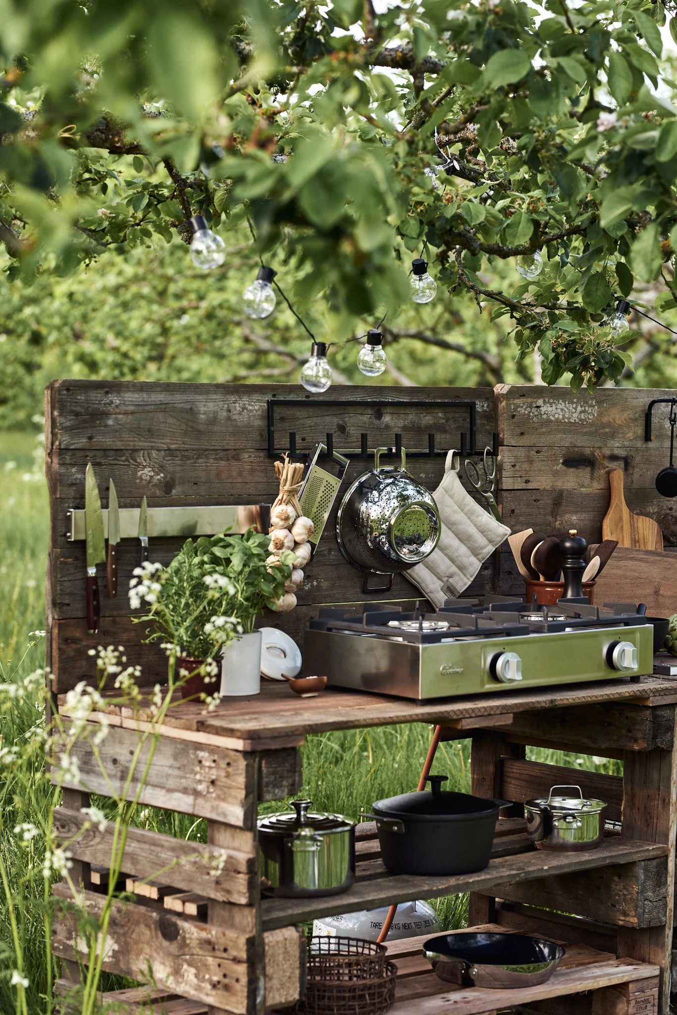 Une cuisine d'extérieur construite à partir de palettes en bois, équipée d'une cuisinière à gaz verte à deux feux, d'ustensiles de cuisine, d'herbes aromatiques et de guirlandes lumineuses.