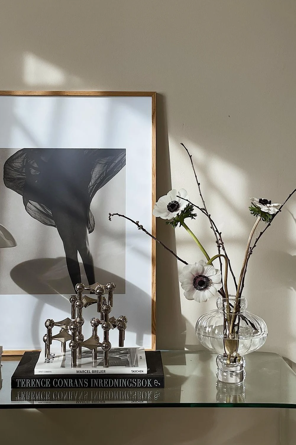 Photo N&B encadrée de jambes avec tissu fluide, bougeoir modulaire argenté, livres et anémones dans un vase.