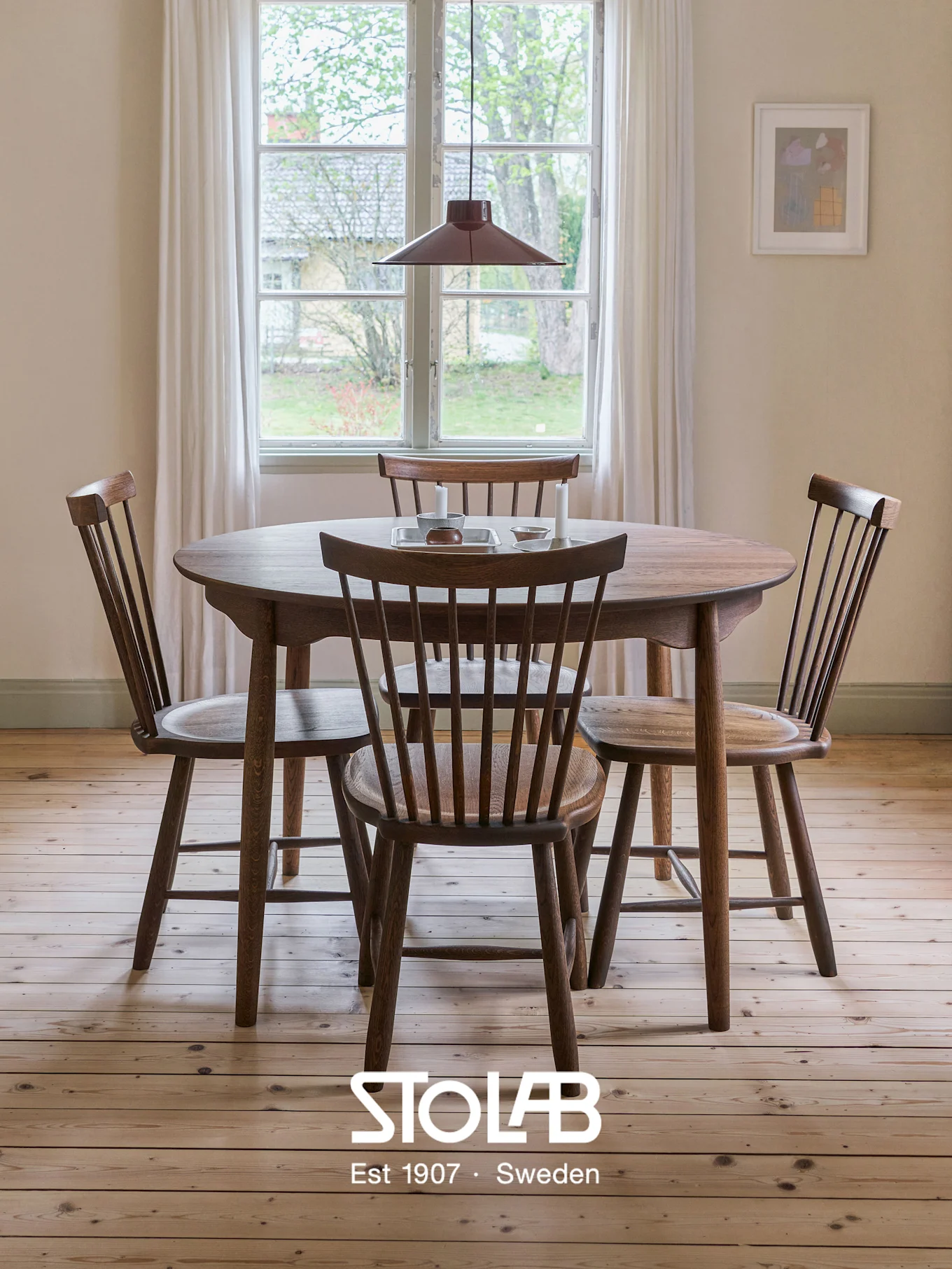 Table à manger ronde en bois avec quatre chaises à dossier à barreaux dans une pièce lumineuse, parquet clair, fenêtre et suspension rouge.
