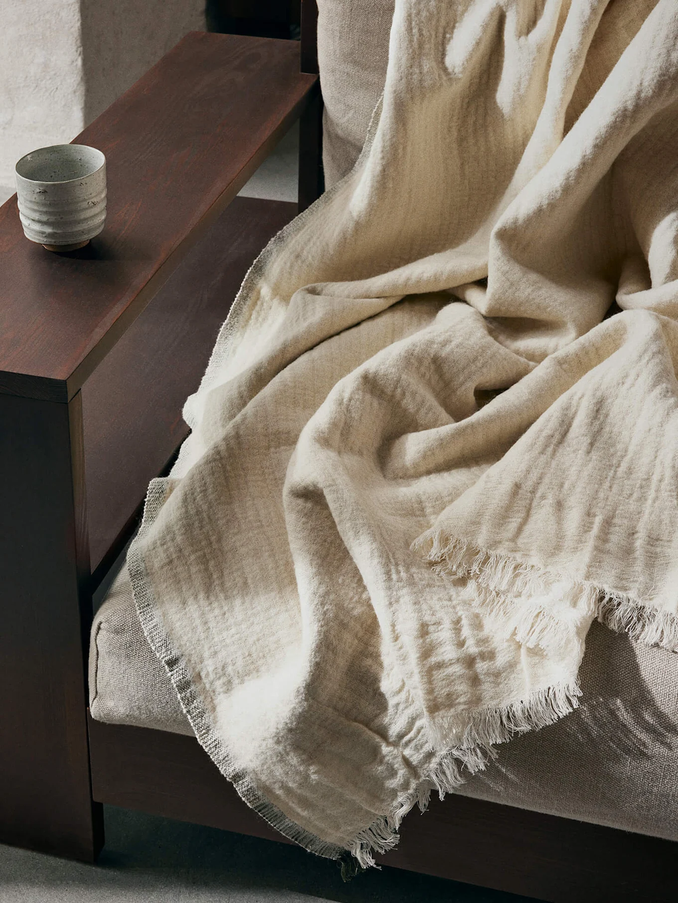 Une couverture texturée beige clair à bords effilochés drapée sur un canapé, avec une tasse en céramique sur un accoudoir en bois foncé.