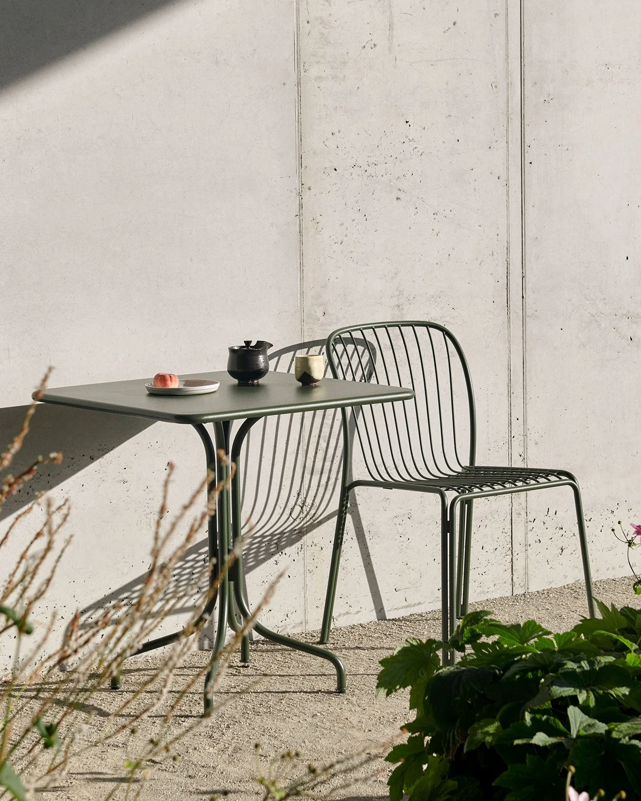 Une table d'extérieur vert foncé avec une pêche, une théière et une tasse, et une chaise à lattes assortie, contre un mur en béton avec de fortes ombres.