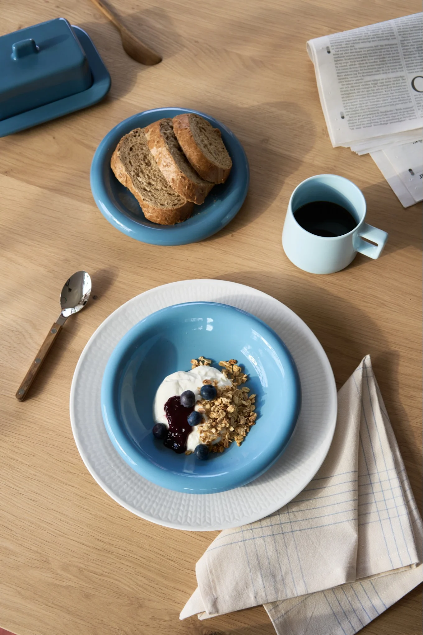 Petit-déjeuner composé de yaourt, de granola, de myrtilles et de pain, servi dans des assiettes bleues et blanches ; café dans une tasse bleue ; corbeille à beurre sur une table en bois.