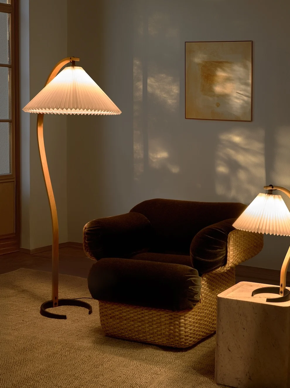 Pièce douillette avec lampadaire incurvé, fauteuil foncé à base tressée et petite lampe de table. La lumière chaude crée des ombres.