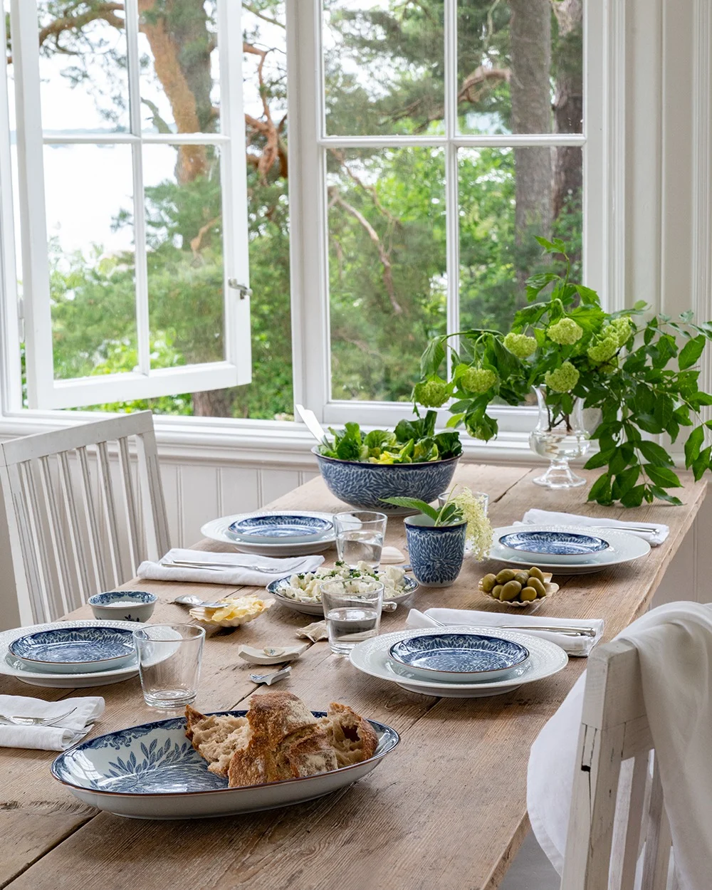 Une table à manger rustique en bois dressée avec de la vaisselle bleue et blanche, de la salade, du pain et des olives, près d'une fenêtre ouverte sur des arbres verts et de l'eau.