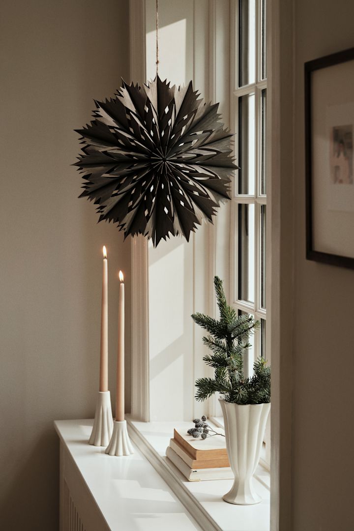 La lanterne en papier en forme d'étoile pour Noël - Venja - de Broste Copenhagen est suspendue à la fenêtre avec une paire de bougeoirs et un vase. 