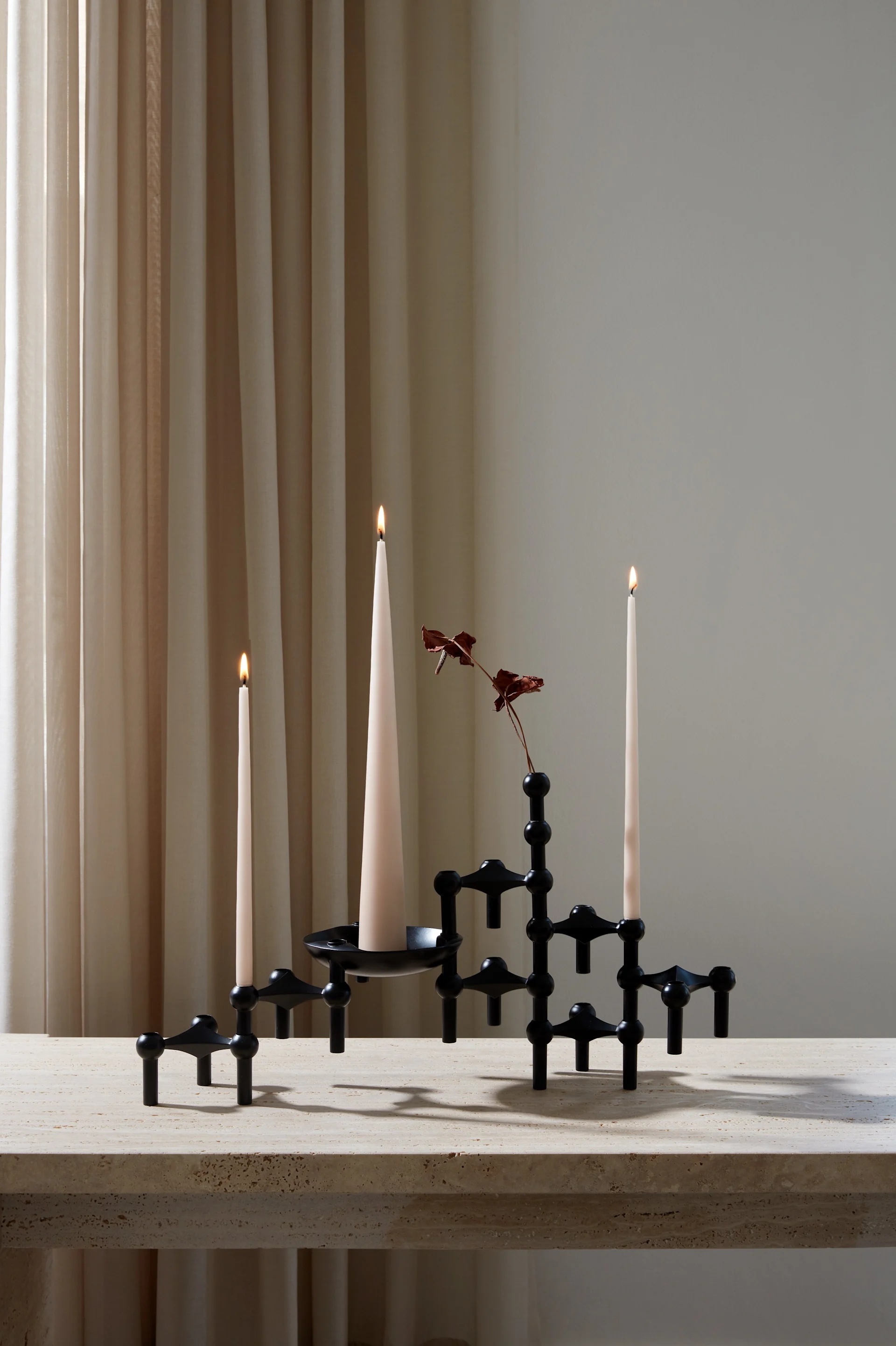Un candélabre modulaire noir avec trois bougies coniques crème allumées et une branche rouge séchée sur une table en pierre beige, devant des rideaux clairs.