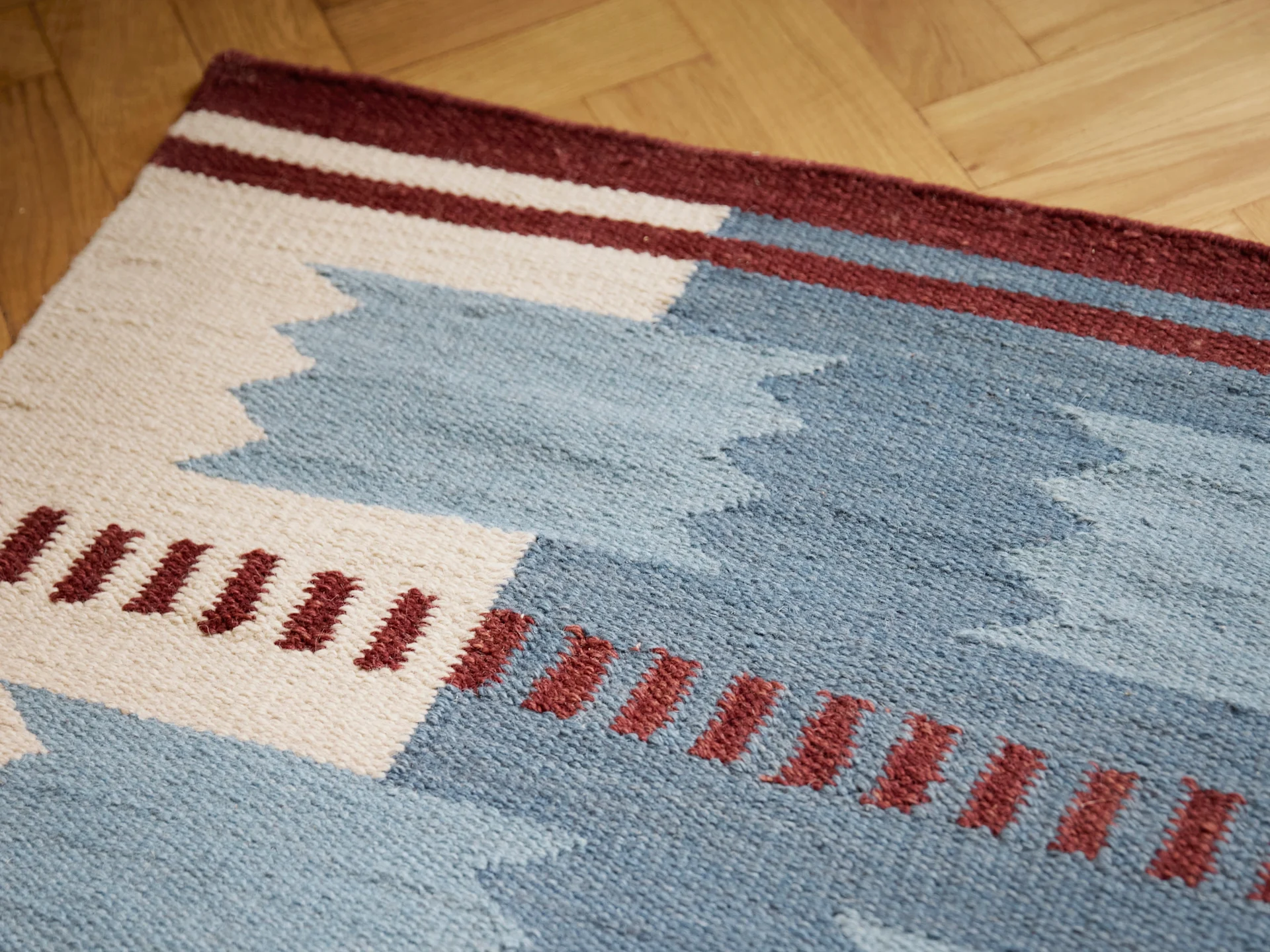 Tapis tissé à motifs géométriques bleus, blancs et bordeaux sur un plancher en bois.