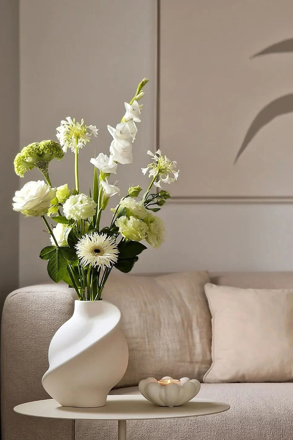 Dans un salon aux tons beiges, une table d’appoint est décorée d’un vase avec des fleurs coupées et d’un photophore.