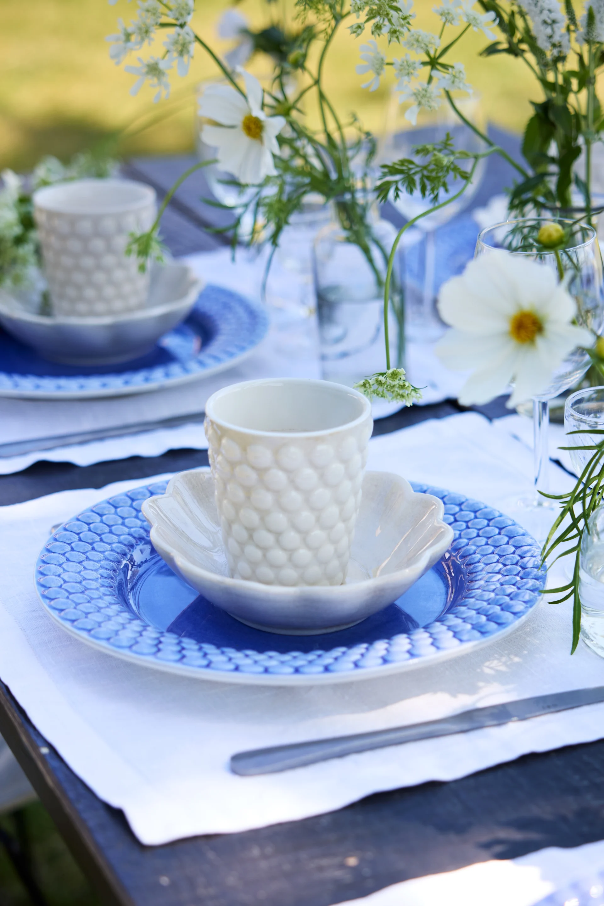 Un gros plan d’une table en plein air dressée en bleu et blanc. Une assiette Bubbles bleue accompagnée d’une tasse Bubbles blanche sur une table décorée de fleurs.