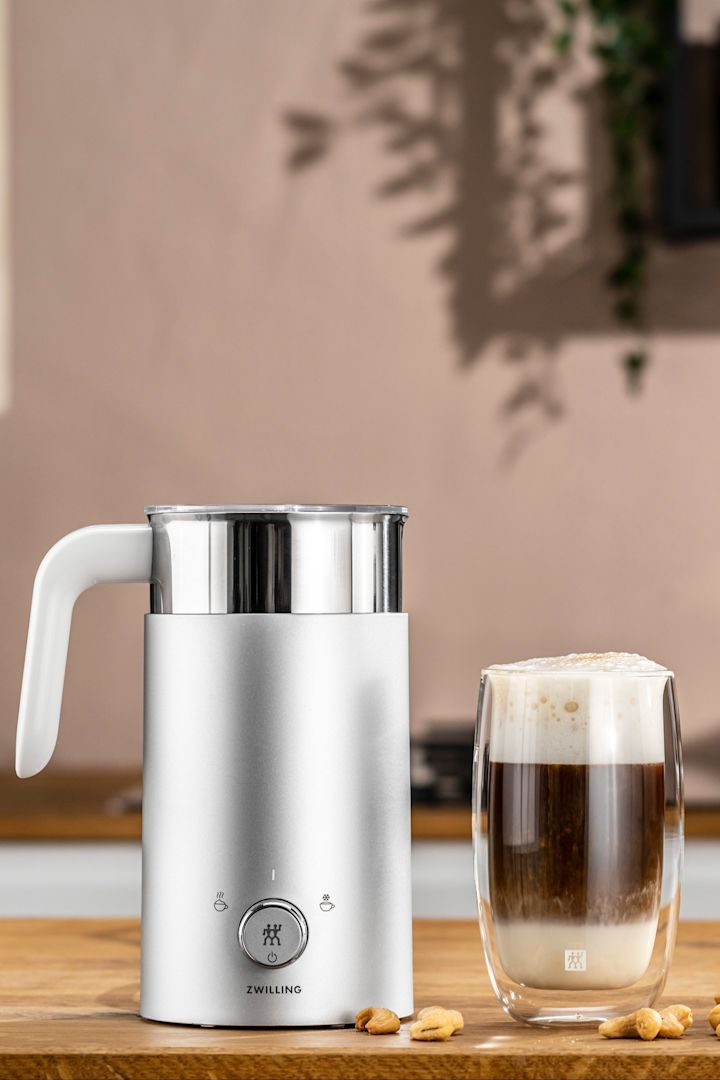 Un mousseur à lait électrique est le cadeau parfait pour les pères qui apprécient un bon cappucchino, car il facilite énormément le travail. Vous voyez ici l'émulsionneur à lait de Zwilling à côté d'une tasse de cappucchino.
