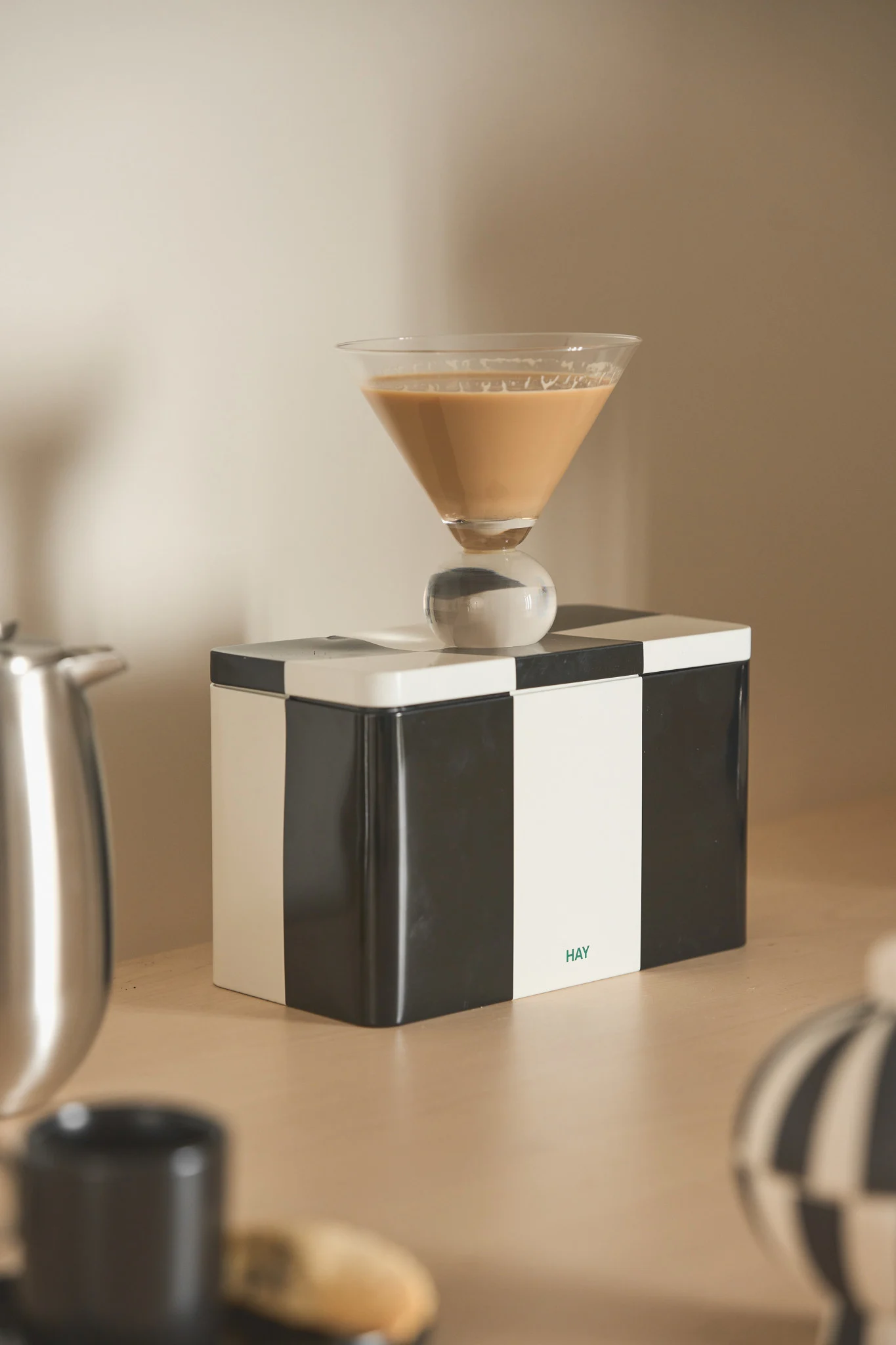 Verre à martini avec boisson marron clair et pied sphérique sur une boîte à carreaux noirs et blancs avec logo 'HAY'.