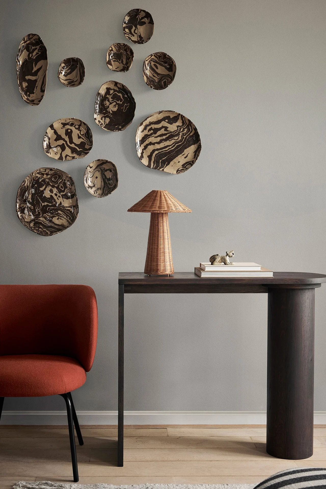 Un mur gris clair orné de plats en céramique marbrés, une console sombre surmontée d'une lampe tressée et une chaise couleur rouille.