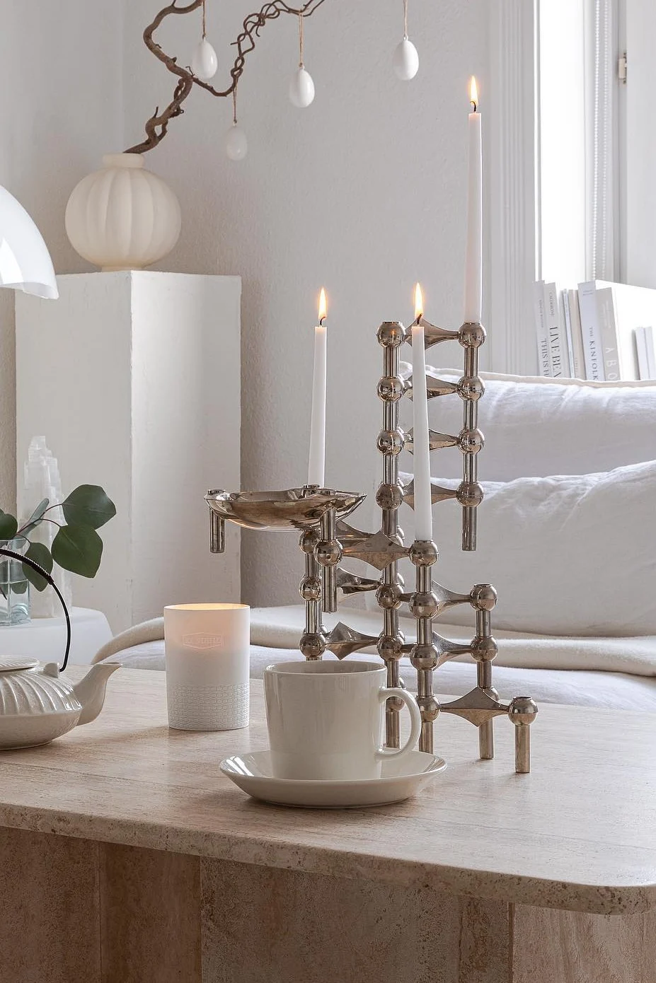 Bougeoir modulaire moderne en argent, orné de trois bougies blanches allumées, d'une tasse à thé et d'une théière, posé sur une table en pierre décorée pour Pâques.