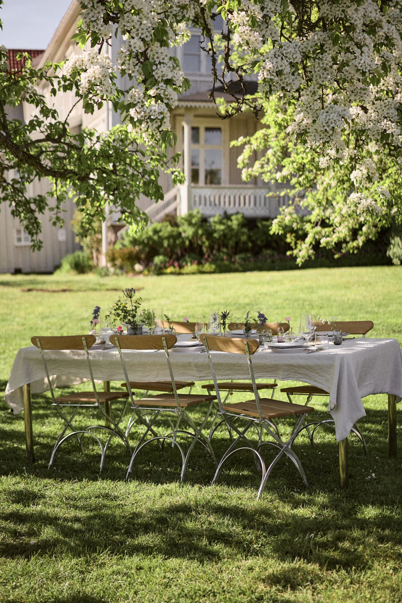 Une table à manger en plein air dressée avec une nappe blanche, des assiettes, des verres et des décorations florales sous un arbre en fleurs dans un jardin ensoleillé.