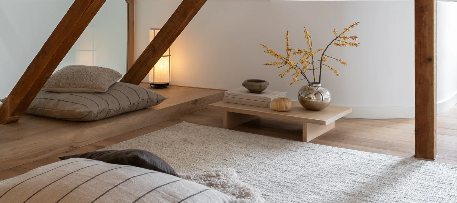Une pièce confortable avec des parquets clairs et des murs blancs, comprenant des coussins de sol, une lampe allumée et une table basse avec un vase argenté contenant des baies jaunes.