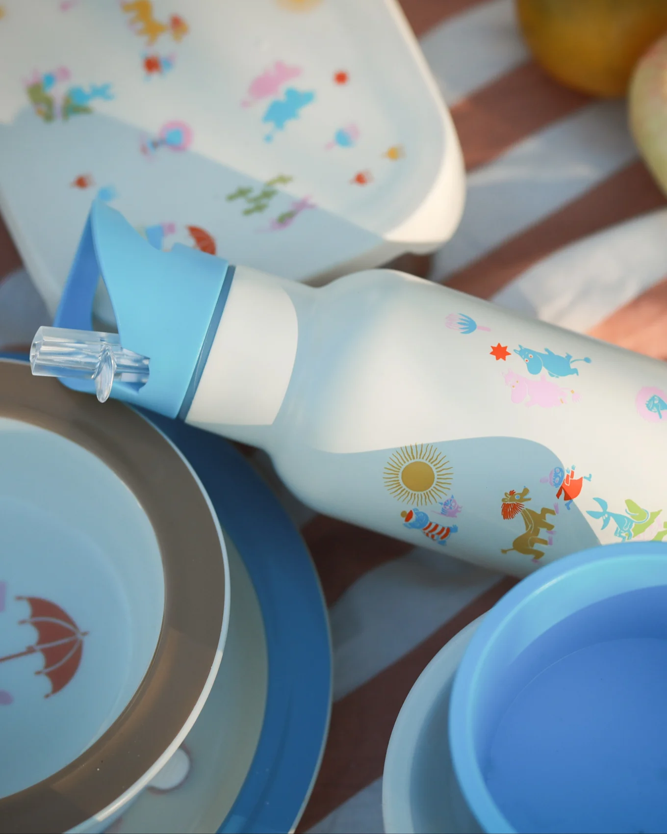 Bouteille d'eau blanche avec paille bleue, personnages de dessins animés colorés et soleil, entourée d'assiettes de pique-nique assorties sur une couverture rayée.