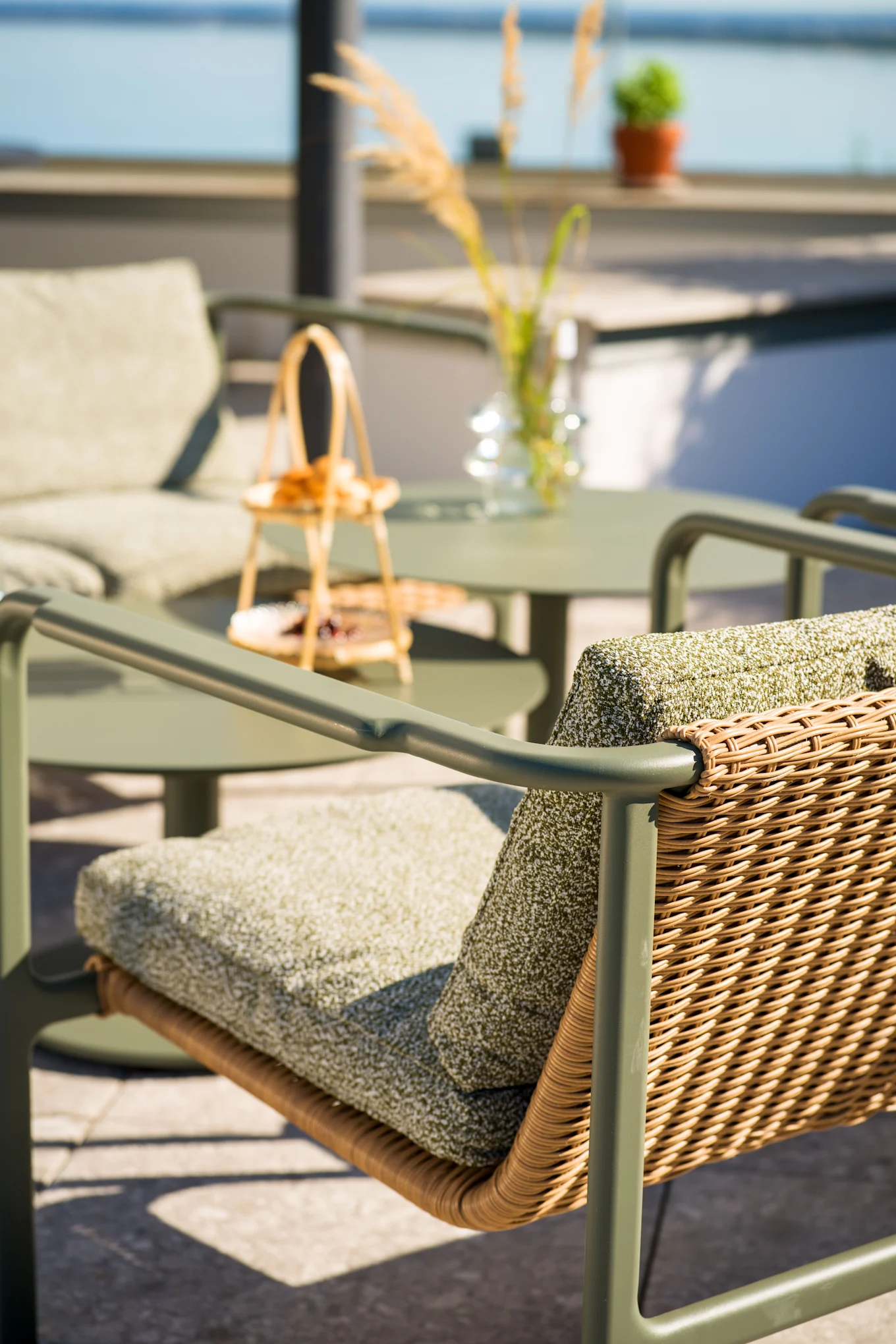 Chaise de patio extérieure avec cadre vert olive, dossier en rotin tressé et coussins verts. Table et chaise assorties en arrière-plan flou près de l'eau.