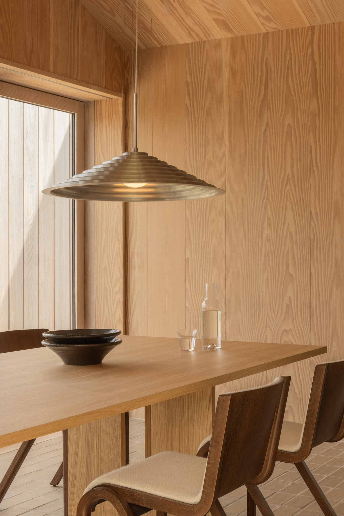 Salle à manger moderne avec murs en bois clair, une table en bois avec deux bols foncés, une bouteille d'eau et une suspension métallique étagée.