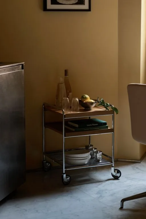 Un chariot de service à trois niveaux avec une structure en métal et des étagères en bois, contenant des bouteilles, des verres, des citrons, des livres et des assiettes.