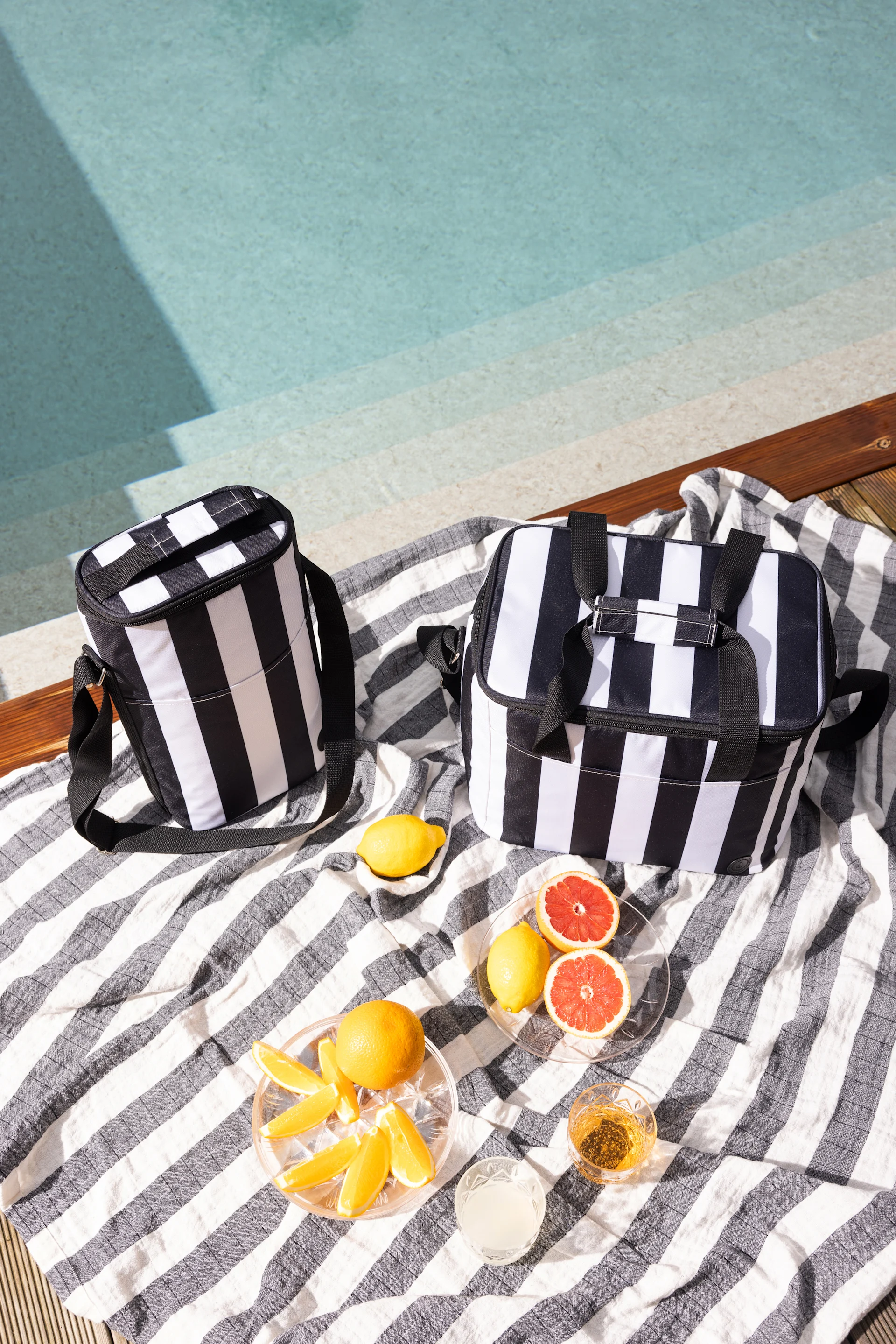 Deux sacs isothermes rayés noir et blanc posés sur une serviette rayée au bord d'une piscine, avec des agrumes et des boissons.