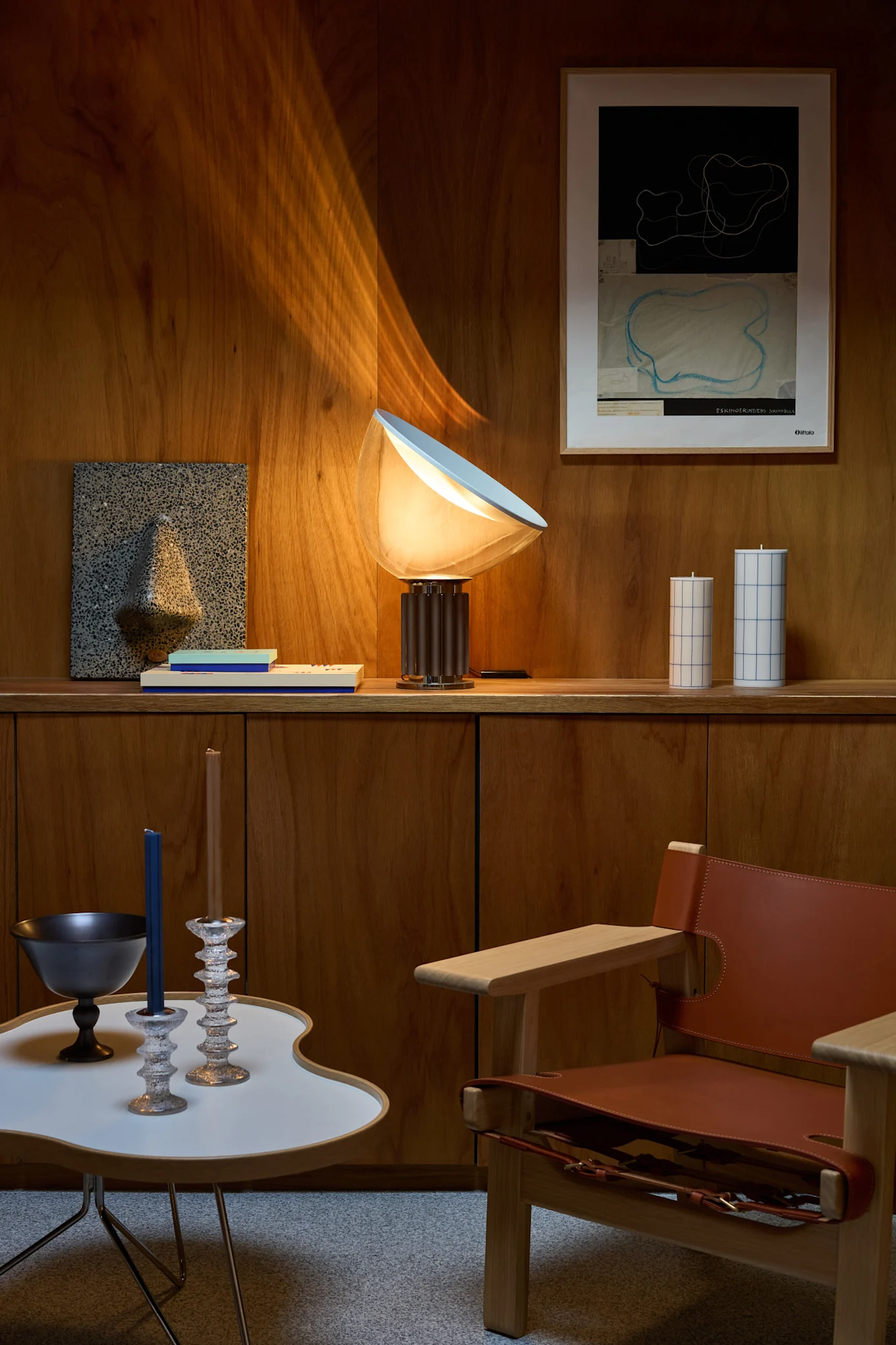 Un espace de vie avec des murs en bois, une lampe de table lumineuse, de l'art mural abstrait, des bougies décoratives, une table d'appoint moderne et un fauteuil en cuir.