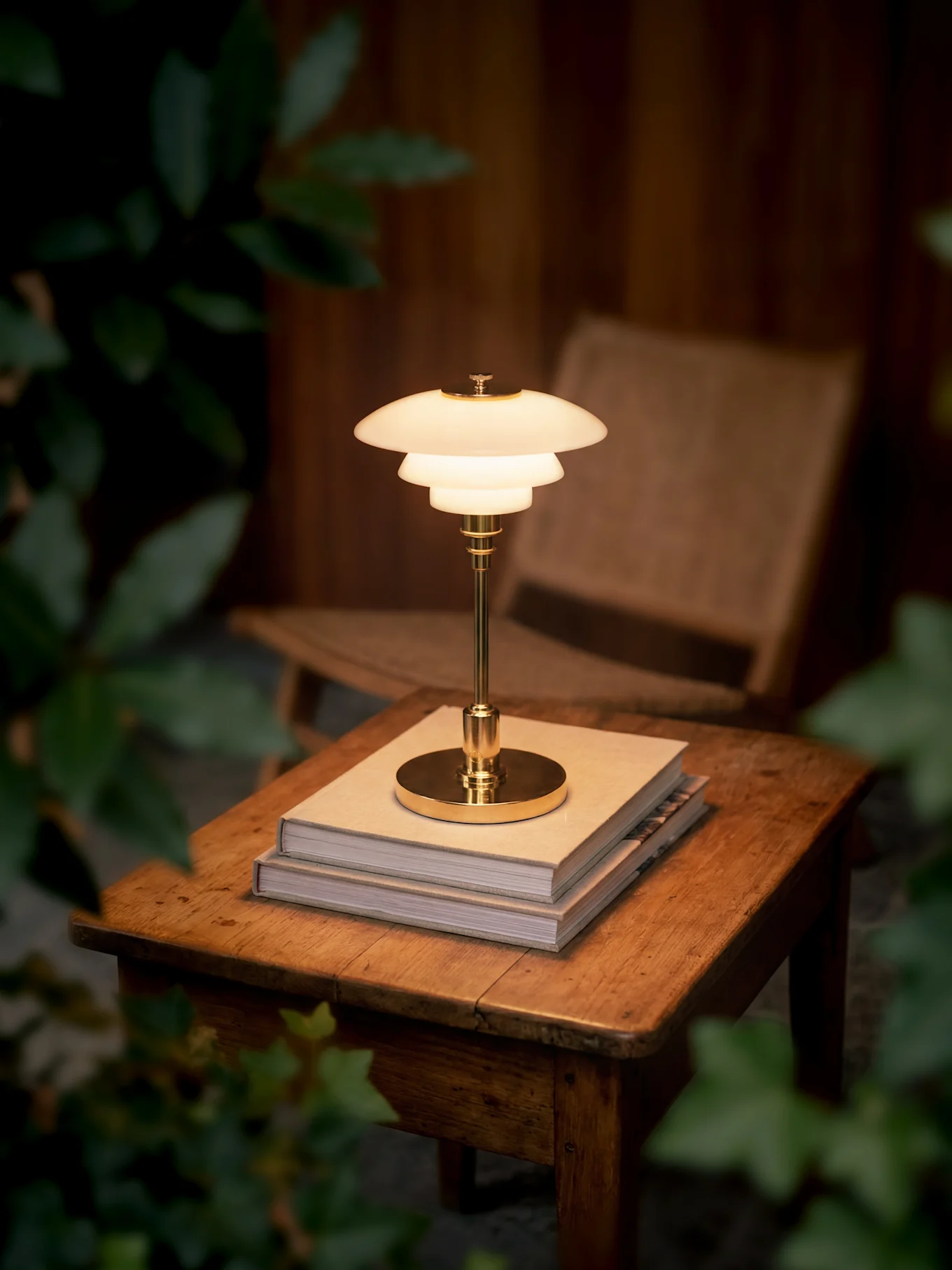 Une lampe de table en laiton brillante à plusieurs niveaux repose sur deux livres sur une table en bois, entourée de feuillage sombre.