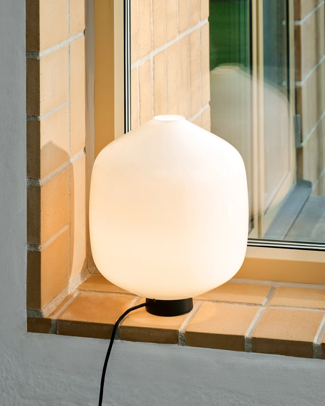 Une lampe de table ronde et blanche lumineuse avec une base et un cordon noirs est posée sur un rebord de fenêtre en briques claires.