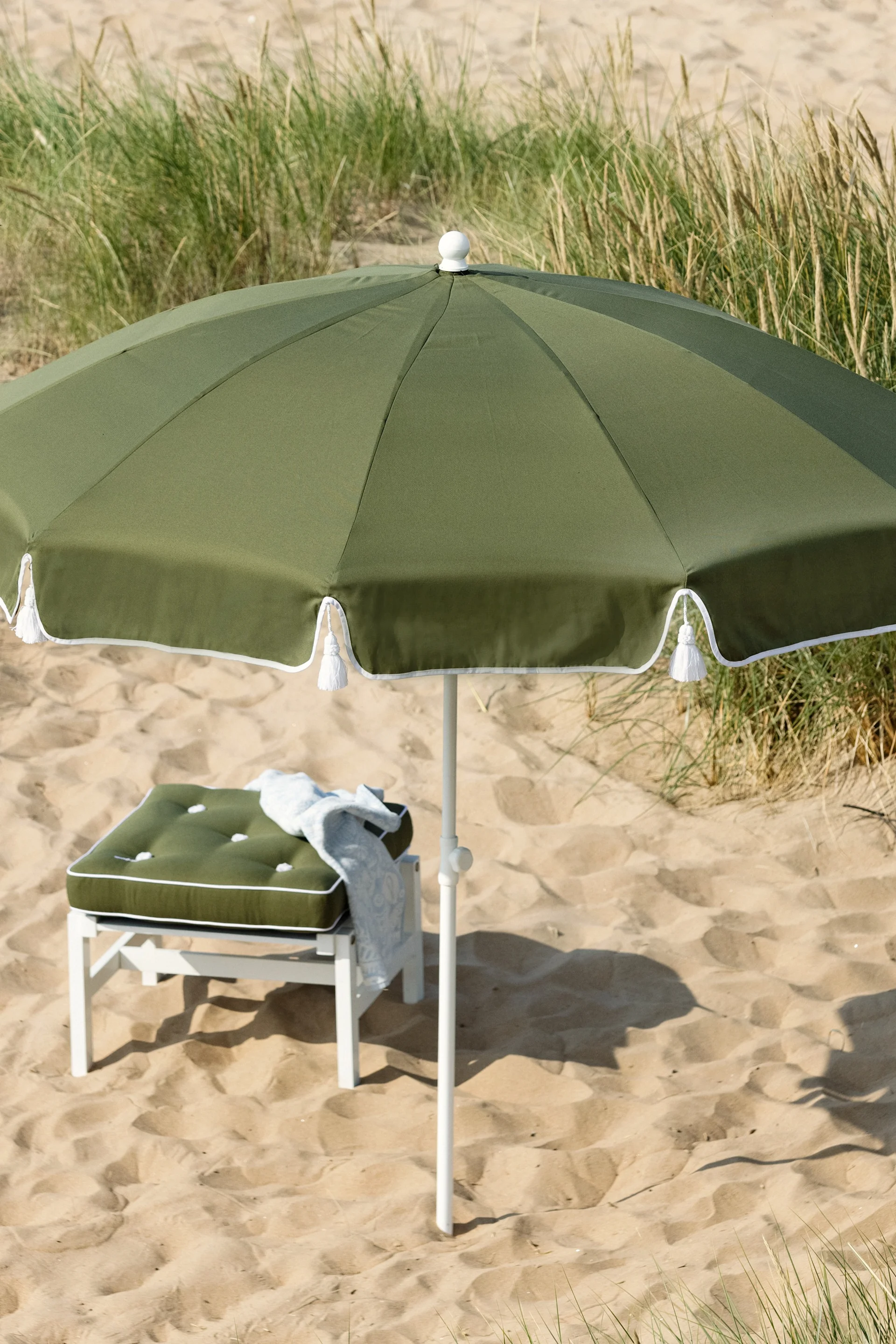 Parasol vert olive et coussin assorti sur un tabouret blanc sur une plage de sable avec de l'herbe.