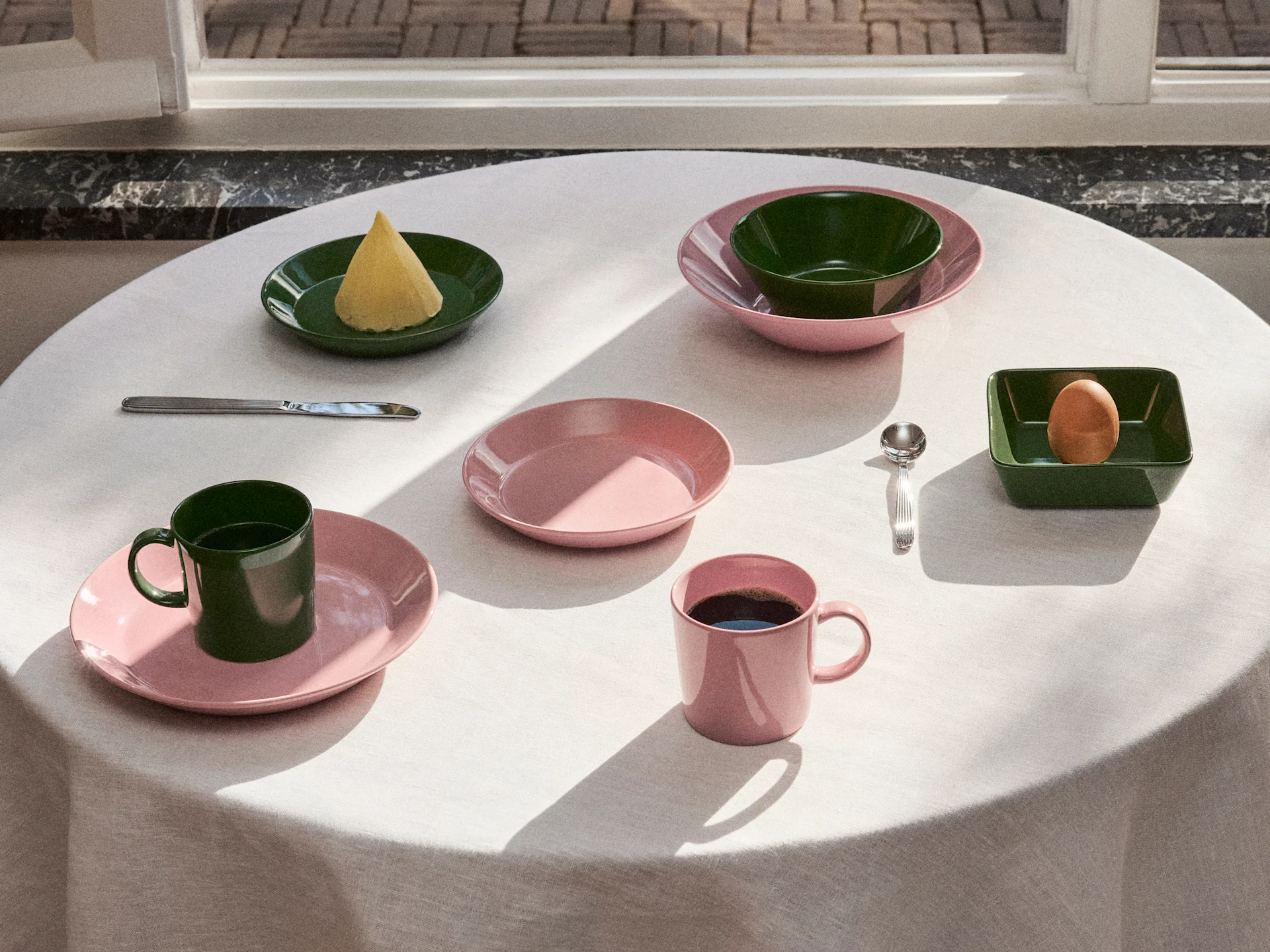 Table dressée avec de la vaisselle rose et vert foncé, assiettes, bols et tasses, avec du beurre, un œuf et du café sous la lumière naturelle.