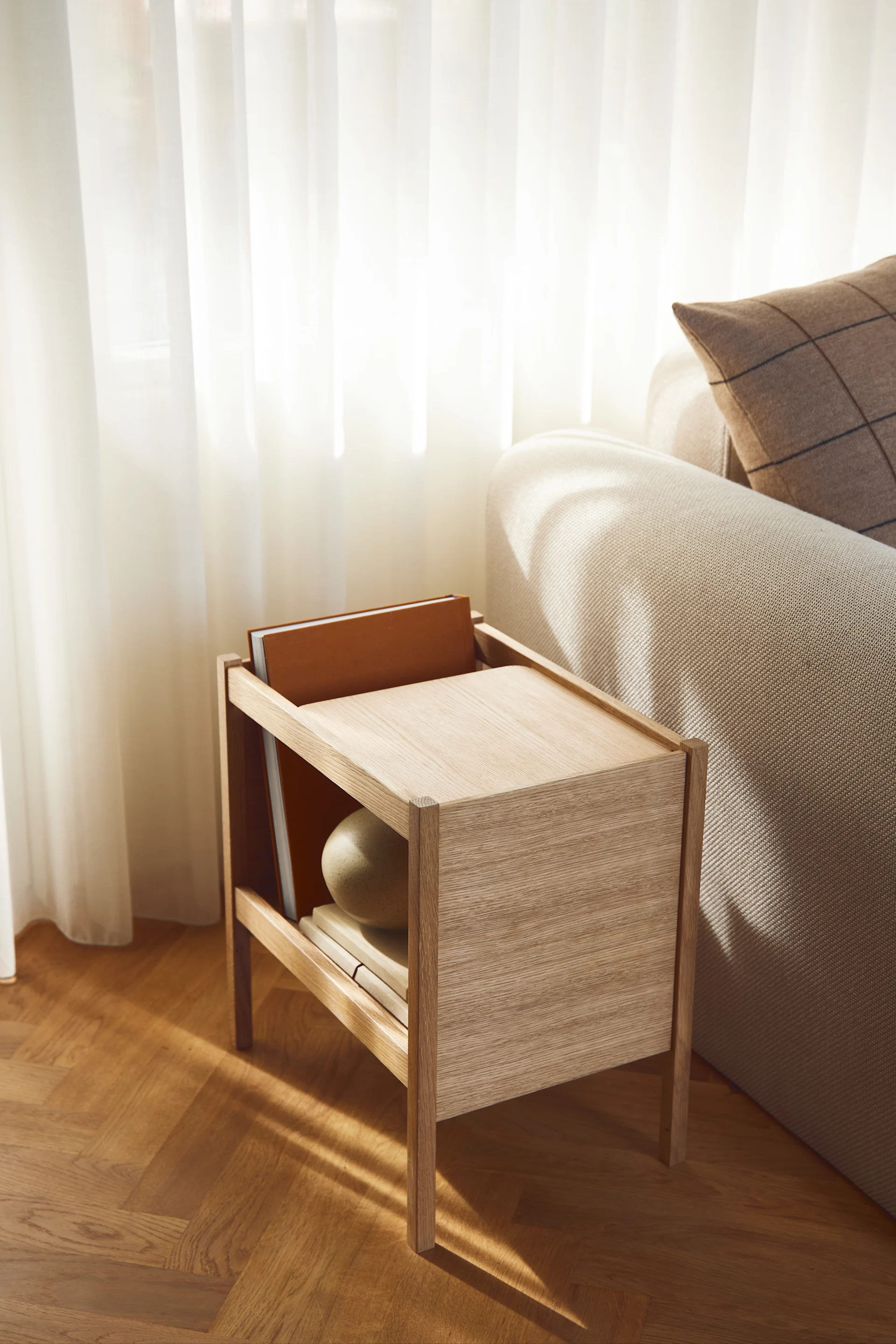 Une table en bois clair sur laquelle sont posés des livres et une boule décorative, à côté d'un canapé beige, baignée par la lumière du soleil qui filtre à travers des rideaux vaporeux.