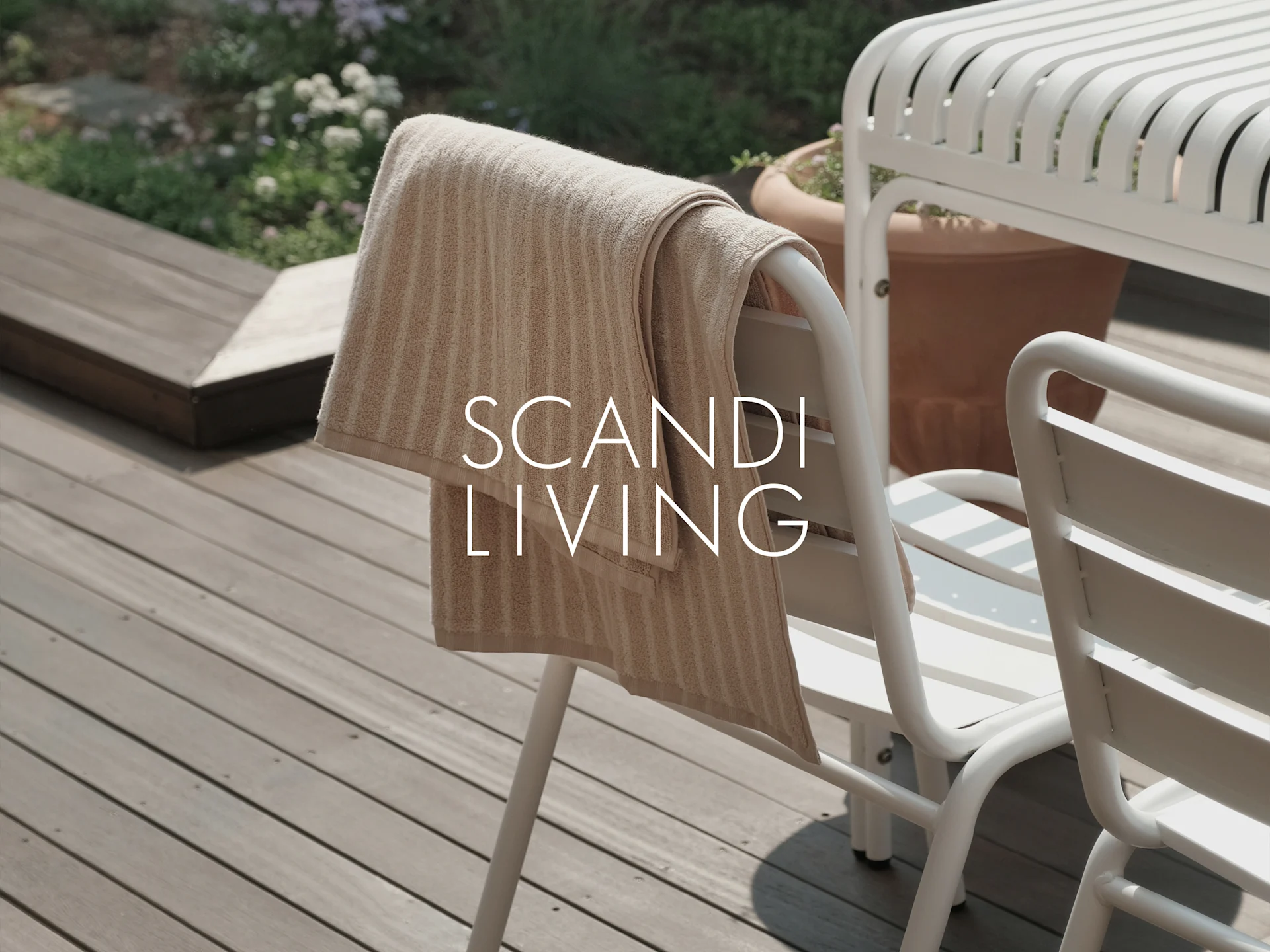 Serviette rayée beige sur chaise d'extérieur blanche sur terrasse en bois avec plantes. Texte: SCANDI LIVING.