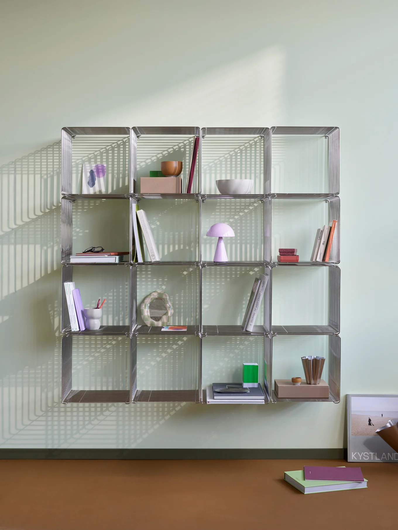Une étagère modulaire chromée avec des livres et des objets décoratifs, éclairée par de fortes ombres linéaires sur un mur vert clair et un sol marron.