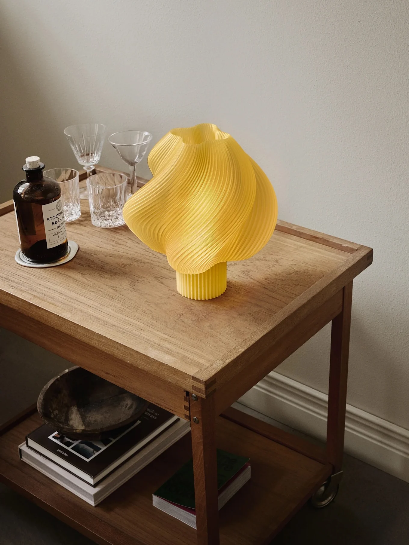 Lampe plissée jaune sur chariot en bois avec bouteille de gin, verres et livres sur l'étagère inférieure.
