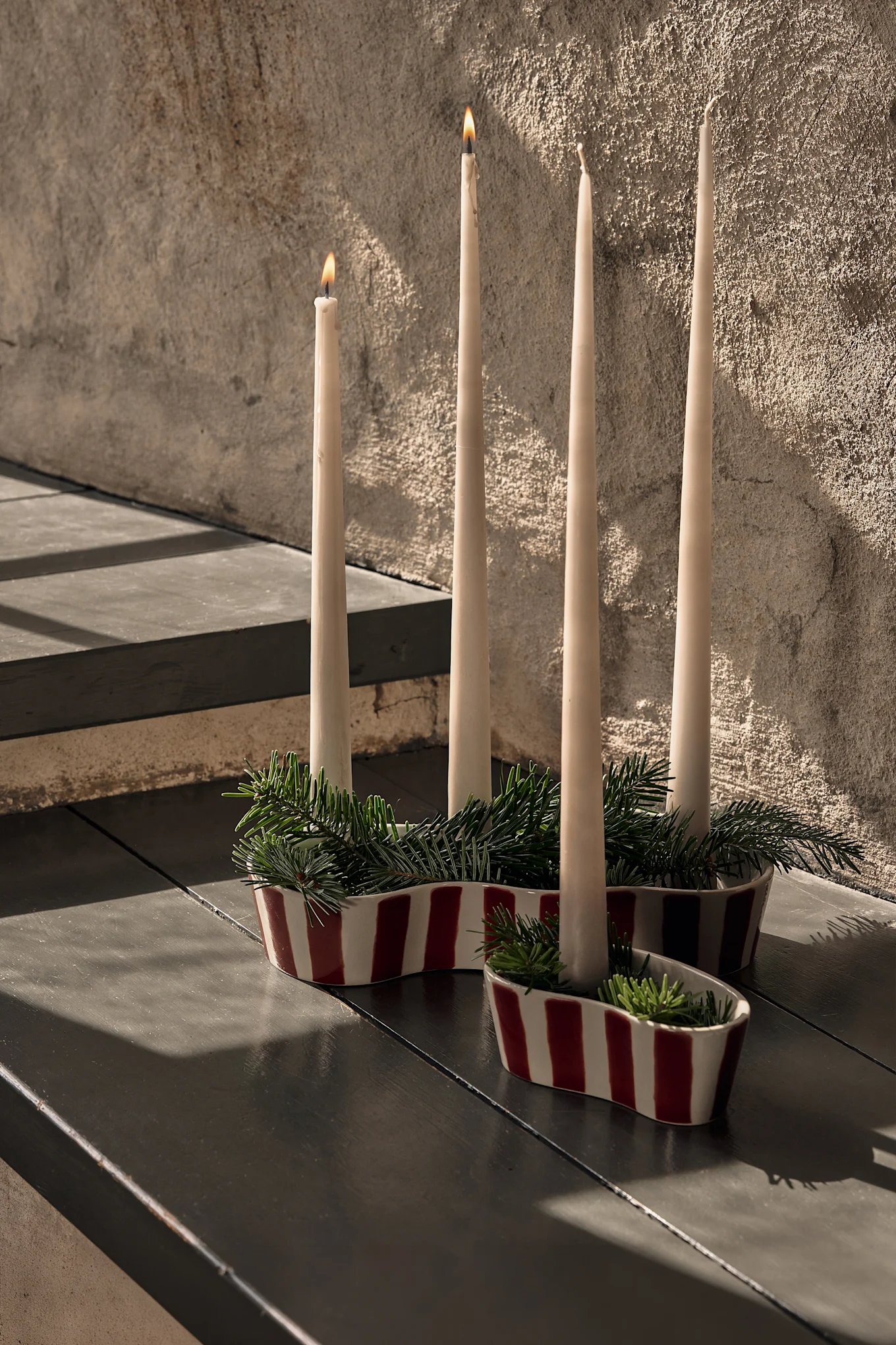 Quatre bougies crème dans des porte-bougies en céramique à rayures rouges et blanches avec des branches de pin, deux bougies allumées, sur une table sombre.
