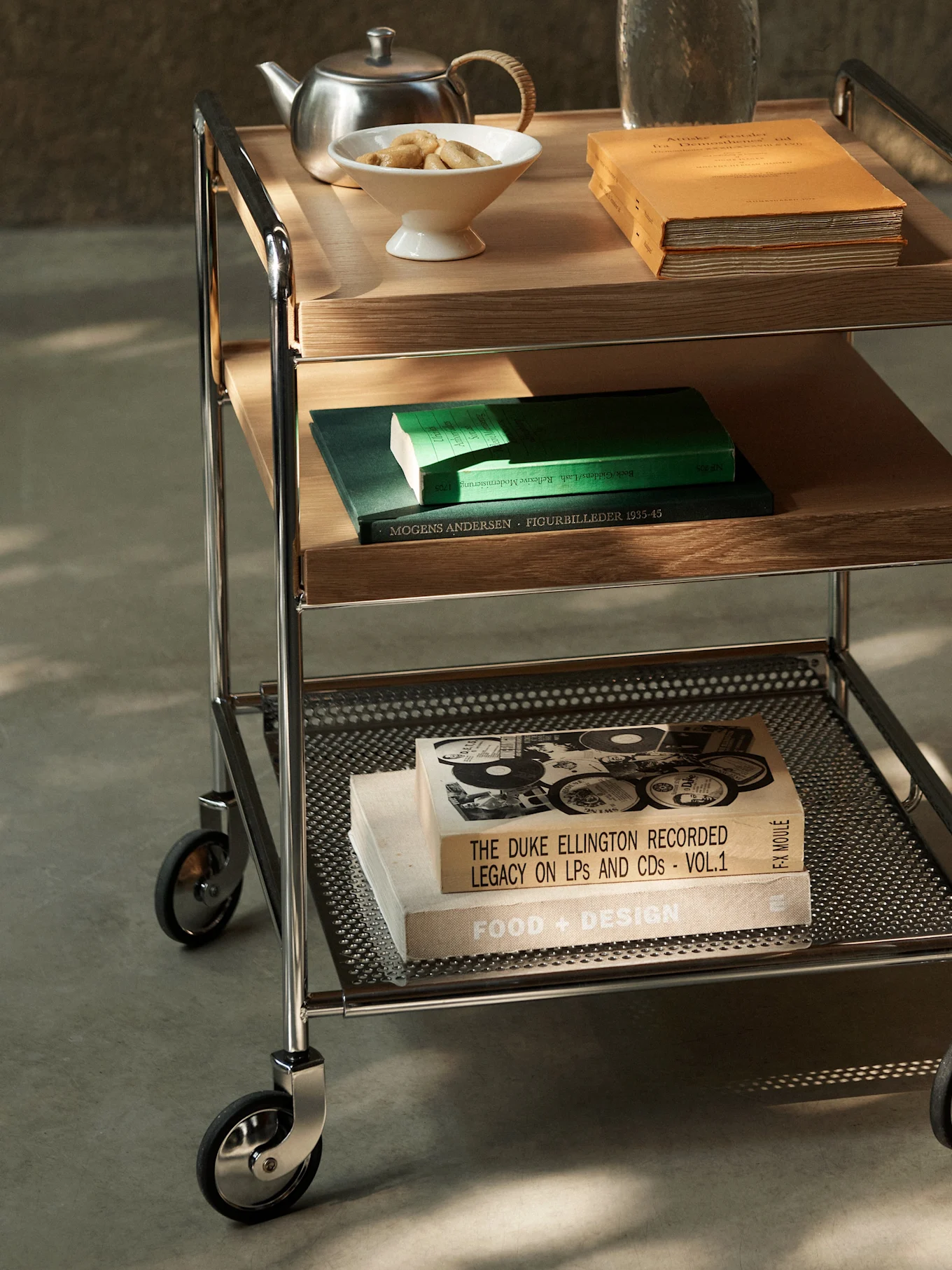 Un chariot de service à trois niveaux avec structure chromée, étagères en bois et métal perforé, contenant des livres, une théière et des biscuits.