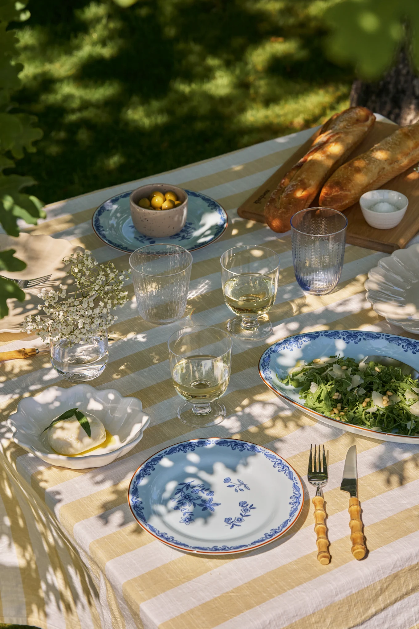 Table à manger extérieure dressée avec nappe rayée blanche et jaune, assiettes florales bleues, vin, salade, mozzarella, olives et baguettes.