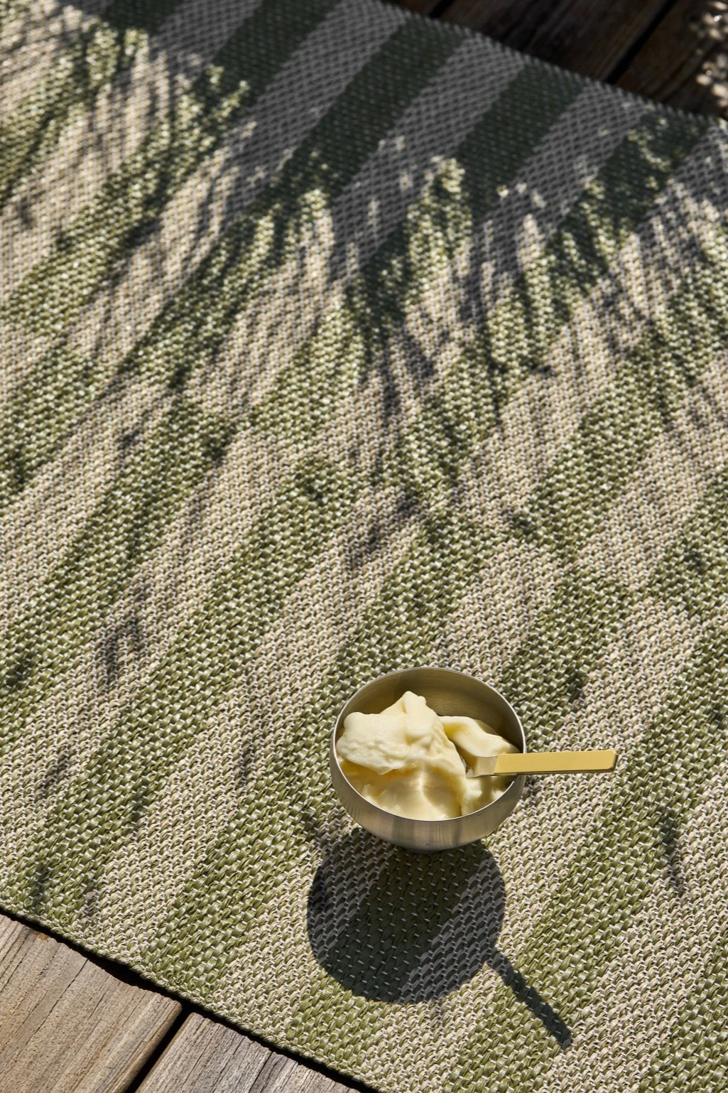 Un bol argenté de glace jaune pâle avec une cuillère dorée repose sur un tapis rayé vert et beige avec des ombres de feuilles, à côté de planches en bois.