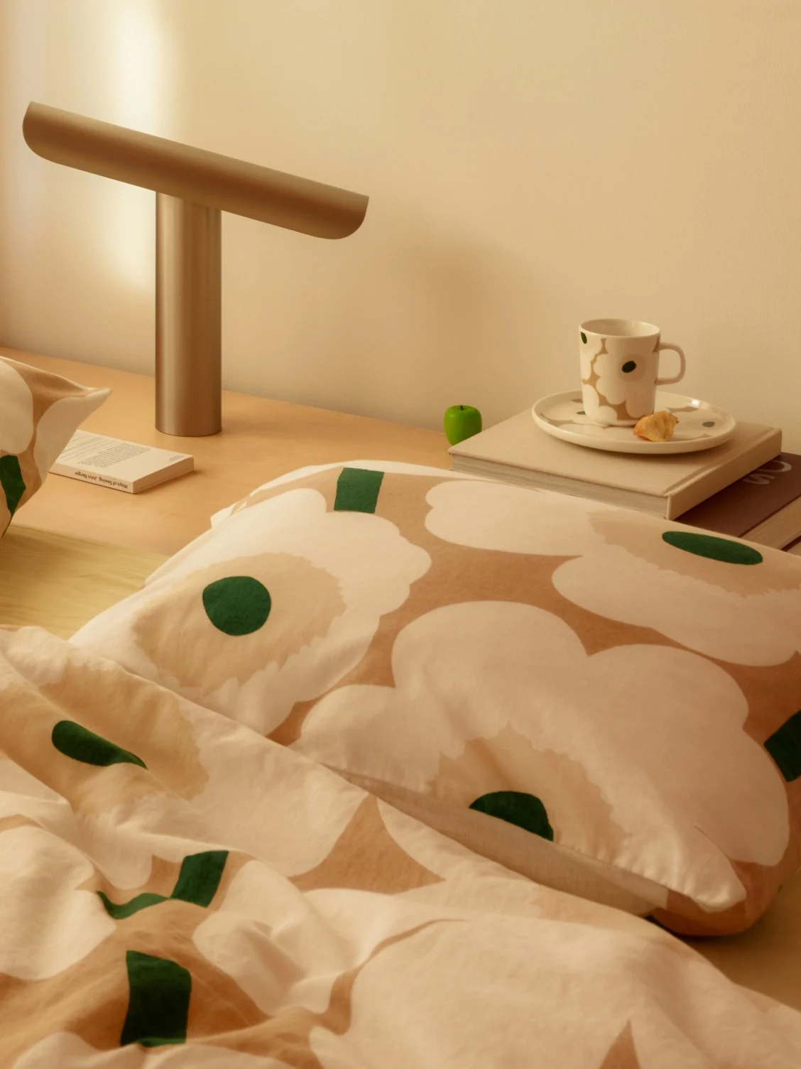Scène de chevet avec une literie florale blanche et beige, une lampe moderne, une tasse et un livre sur une table de chevet en bois.