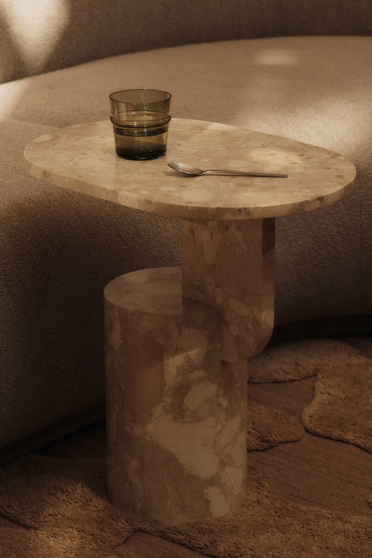 Une table d'appoint en marbre beige sur laquelle sont posés un verre d'eau vert foncé et une cuillère en argent, à côté d'un canapé et d'un tapis à la texture marquée.