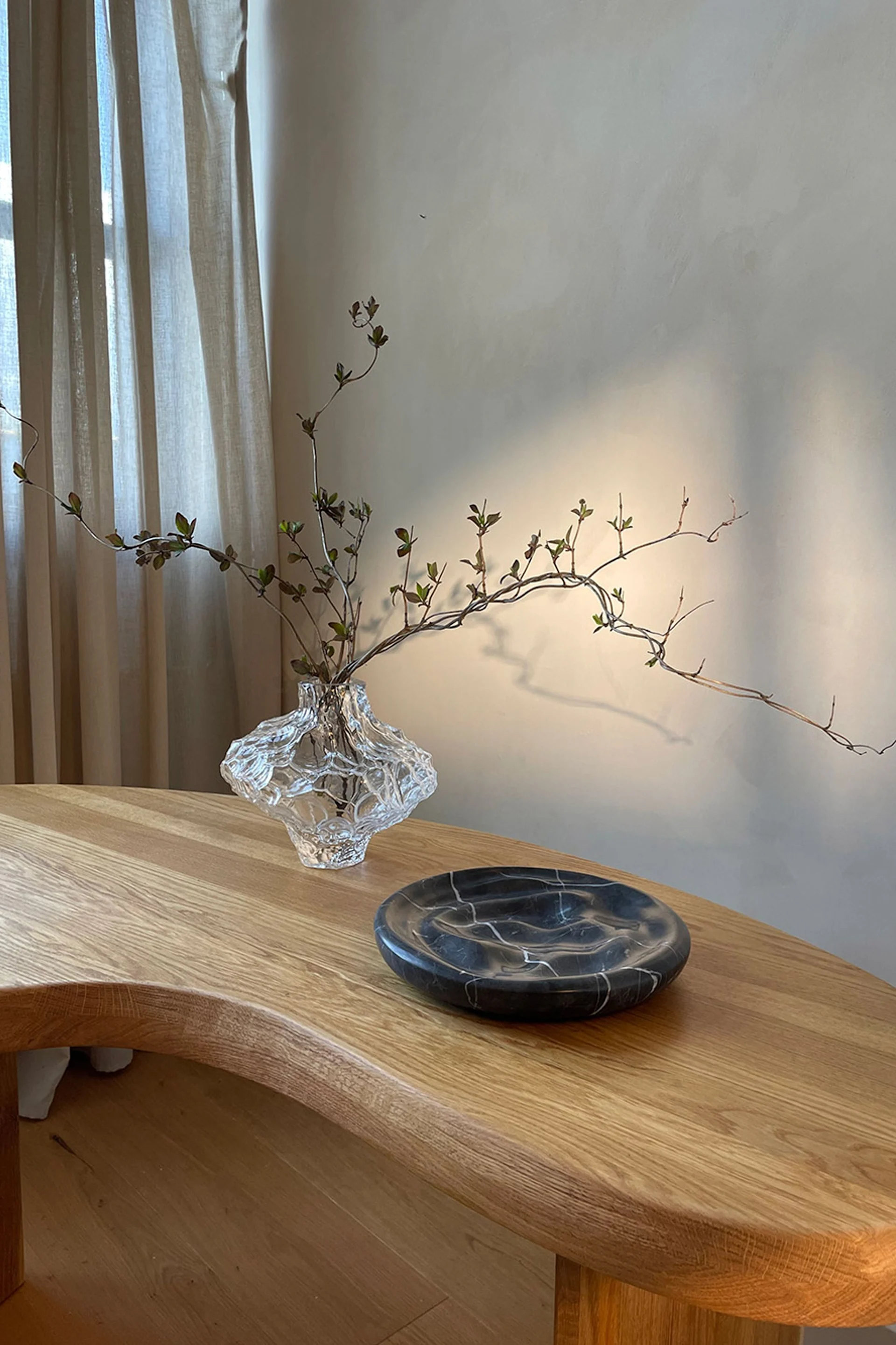 Vase en verre texturé transparent avec des branches feuillues et une assiette en marbre foncé sur une table en bois clair.