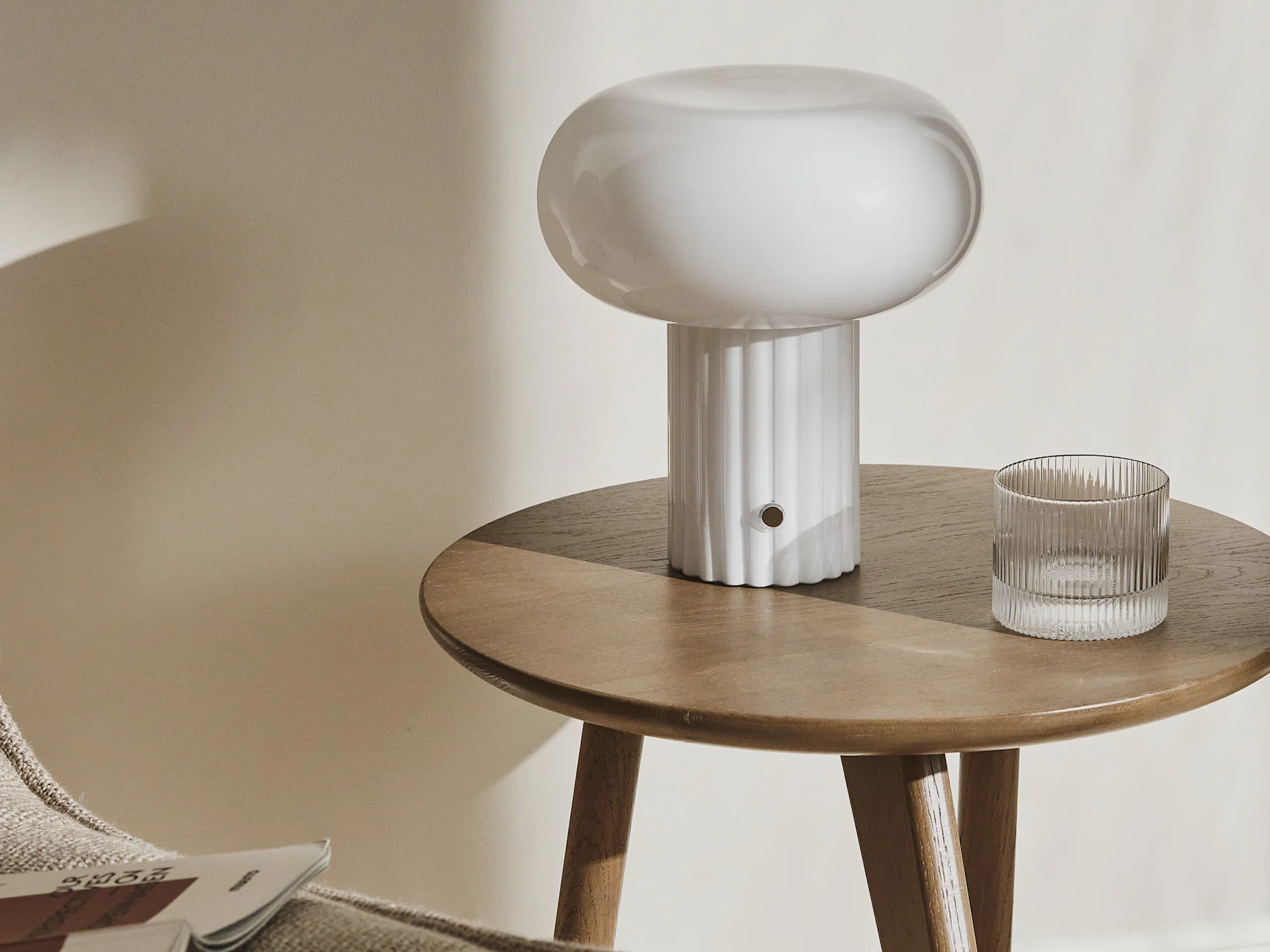 Une lampe champignon blanche et un verre côtelé sur une table d'appoint ronde en bois.