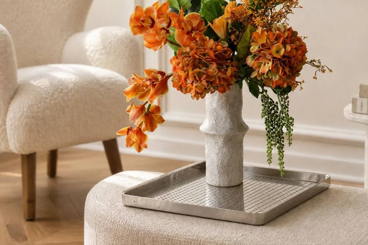 Fleurs orange éclatantes dans un vase blanc texturé sur un plateau argenté, posées sur un pouf clair dans un salon moderne.