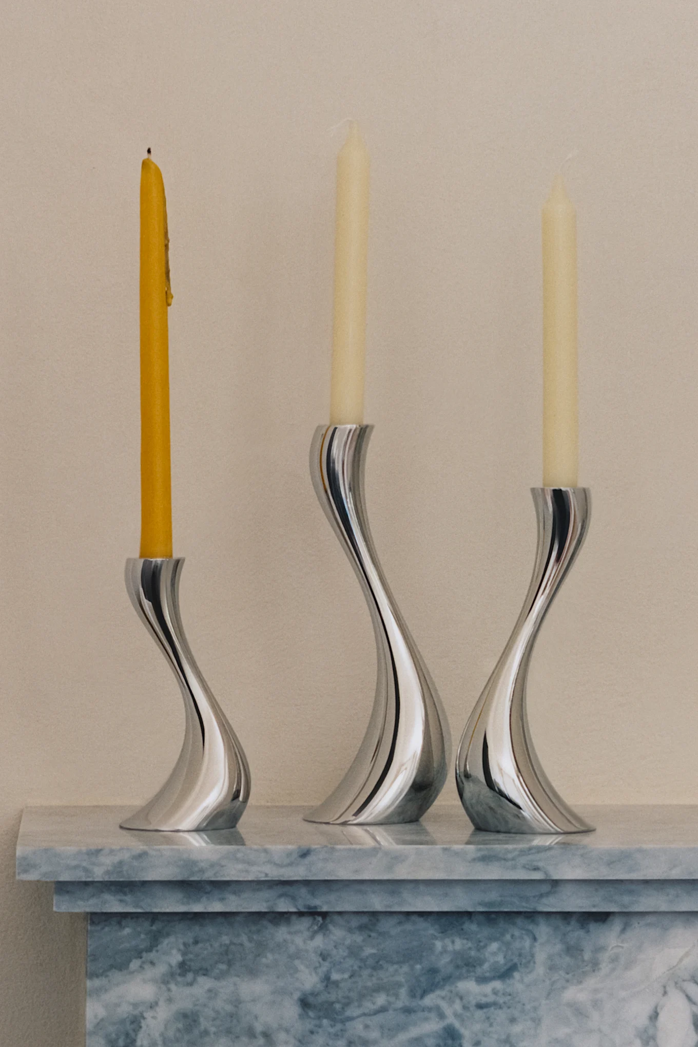 Trois bougeoirs élégants, argentés et courbés avec une bougie jaune brûlée et deux bougies crème non allumées sur une surface en marbre.