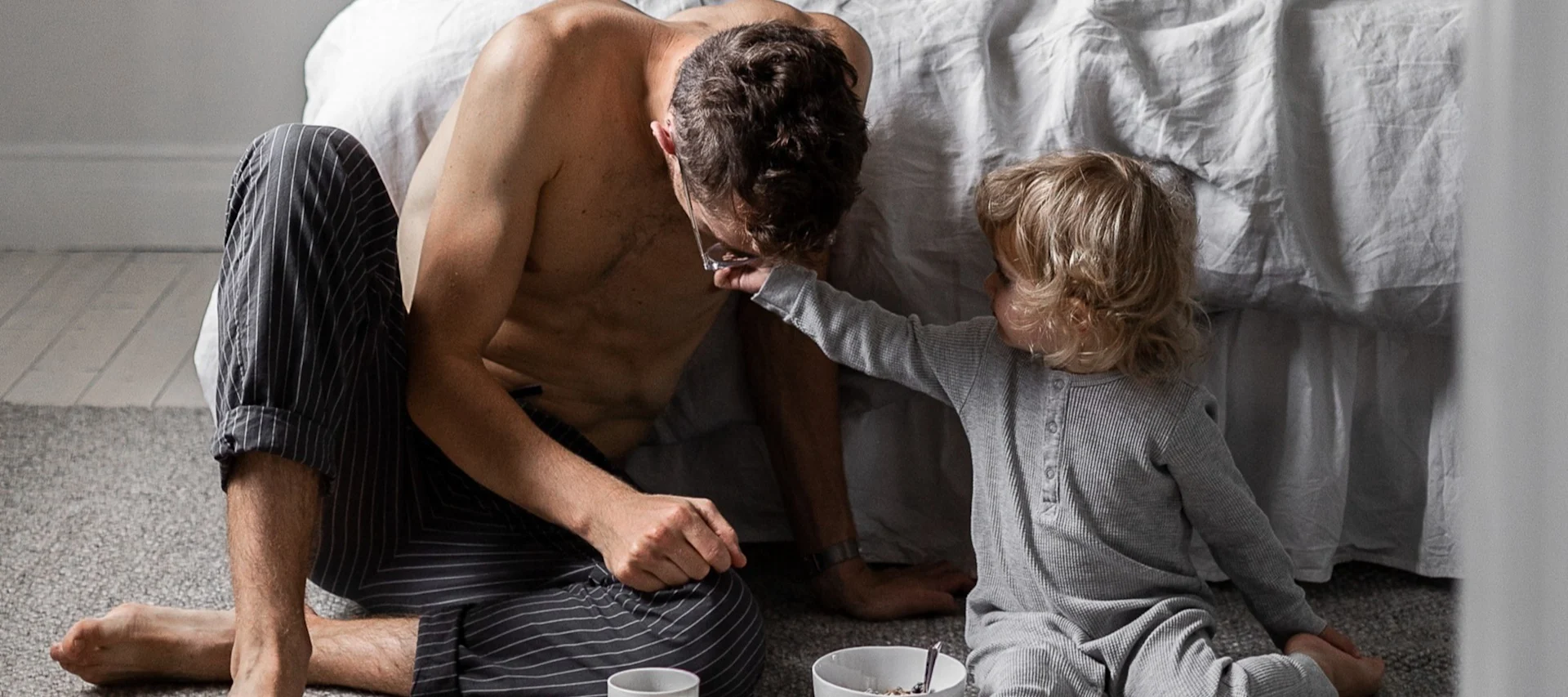 Père et enfant assis sur le sol de la chambre, l'enfant touchant les lunettes du père, petit-déjeuner à proximité.